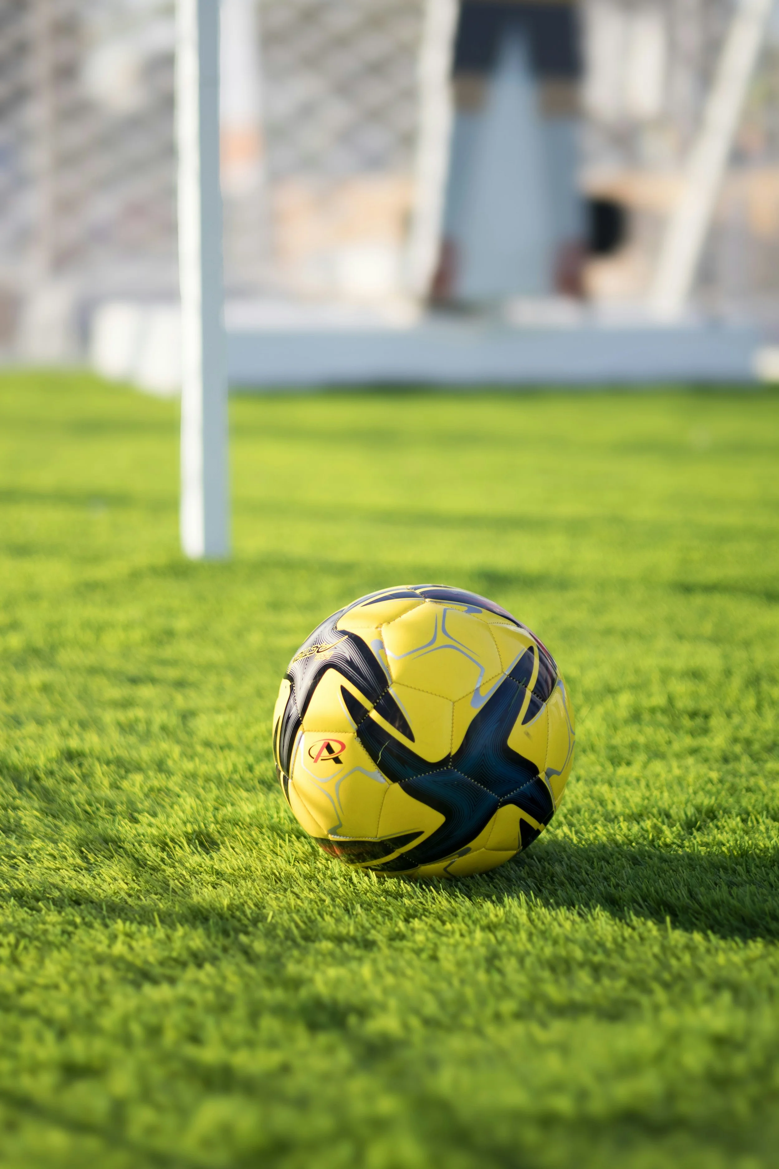 Close-up of a black and yellow soccer ball on a grassy field with goalposts and a person in the background.