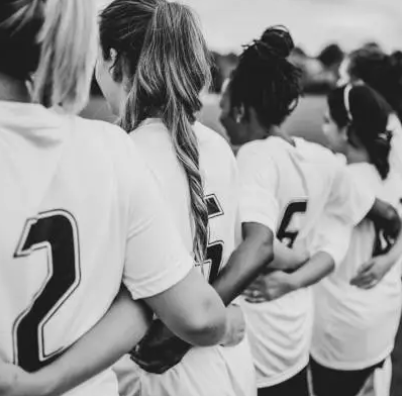 A group of female soccer players standing in a line, arm in arm, outdoors.
