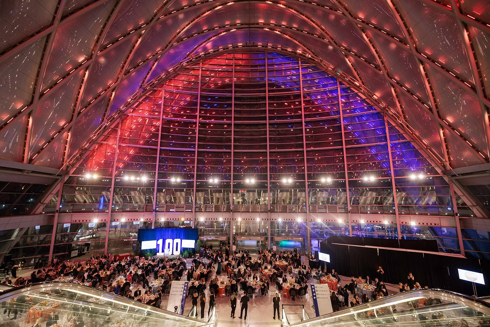 Interior of a large modern building with a high, arched glass ceiling illuminated in pink, purple, and blue lights. Numerous people are seated at round tables for a formal event, with screens and signage visible.