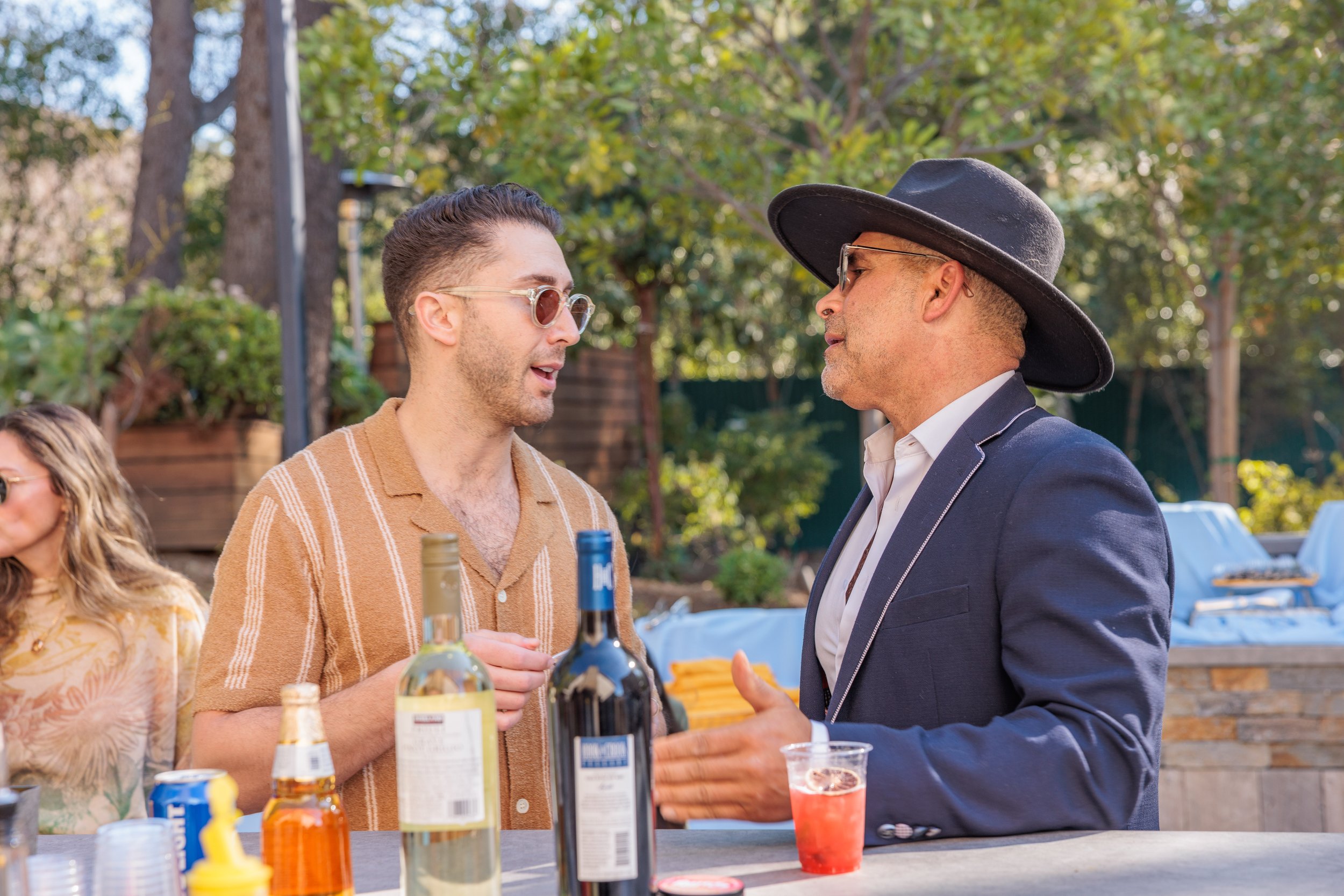Two men conversing at an outdoor gathering with drinks on the table. One man has sunglasses and wears a beige shirt, the other man wears a black wide-brimmed hat and a navy blazer.