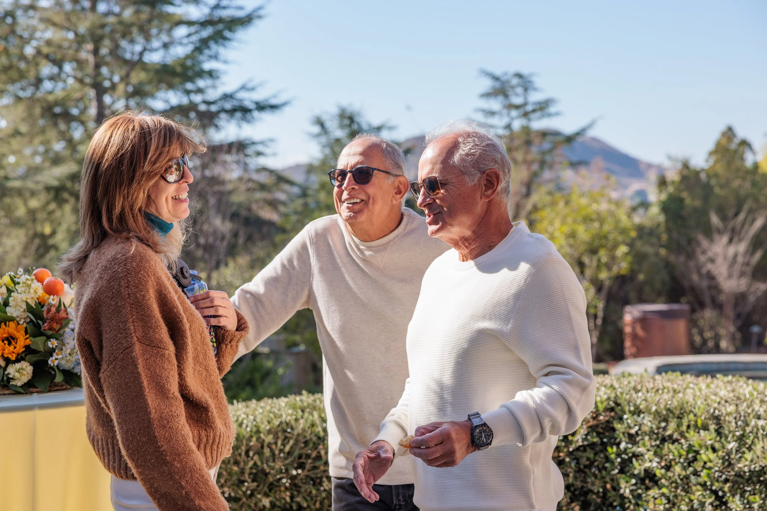 Three elderly adults outdoors, smiling and talking, with trees and mountains in the background.