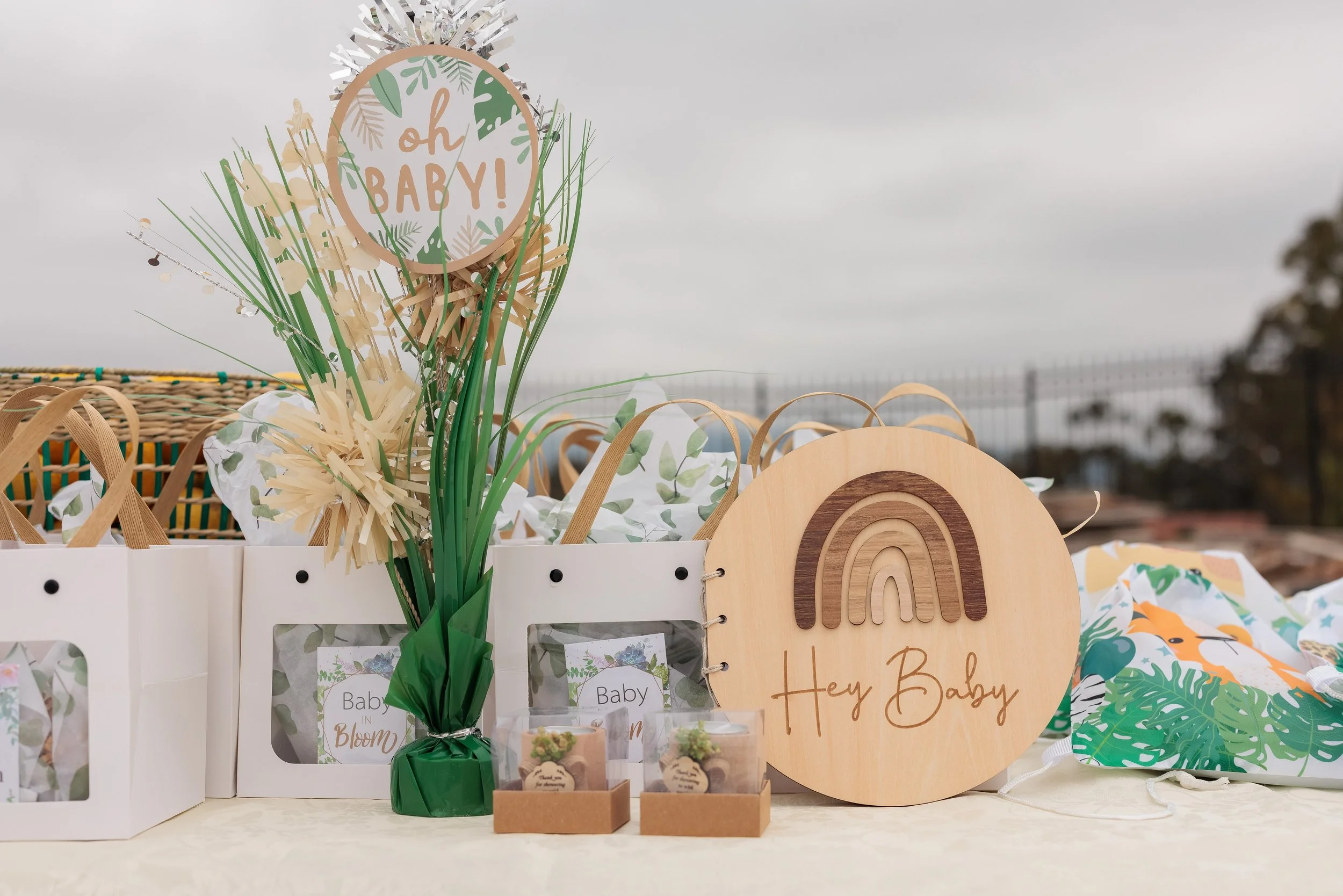 Decorative baby shower display with a sign saying 'oh BABY!', a wooden sign saying 'Hey Baby' with a rainbow, and various gift items, including gift bags and small boxes, on a table outdoors with a cloudy sky.