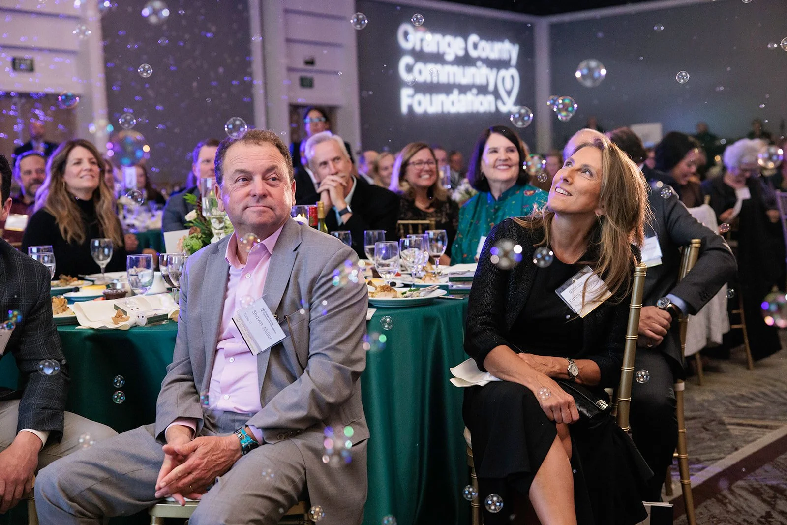 People attending a formal event, seated at tables with green tablecloths, surrounded by floating bubbles, and a large screen displaying the logo for Orange County Community Foundation in the background.