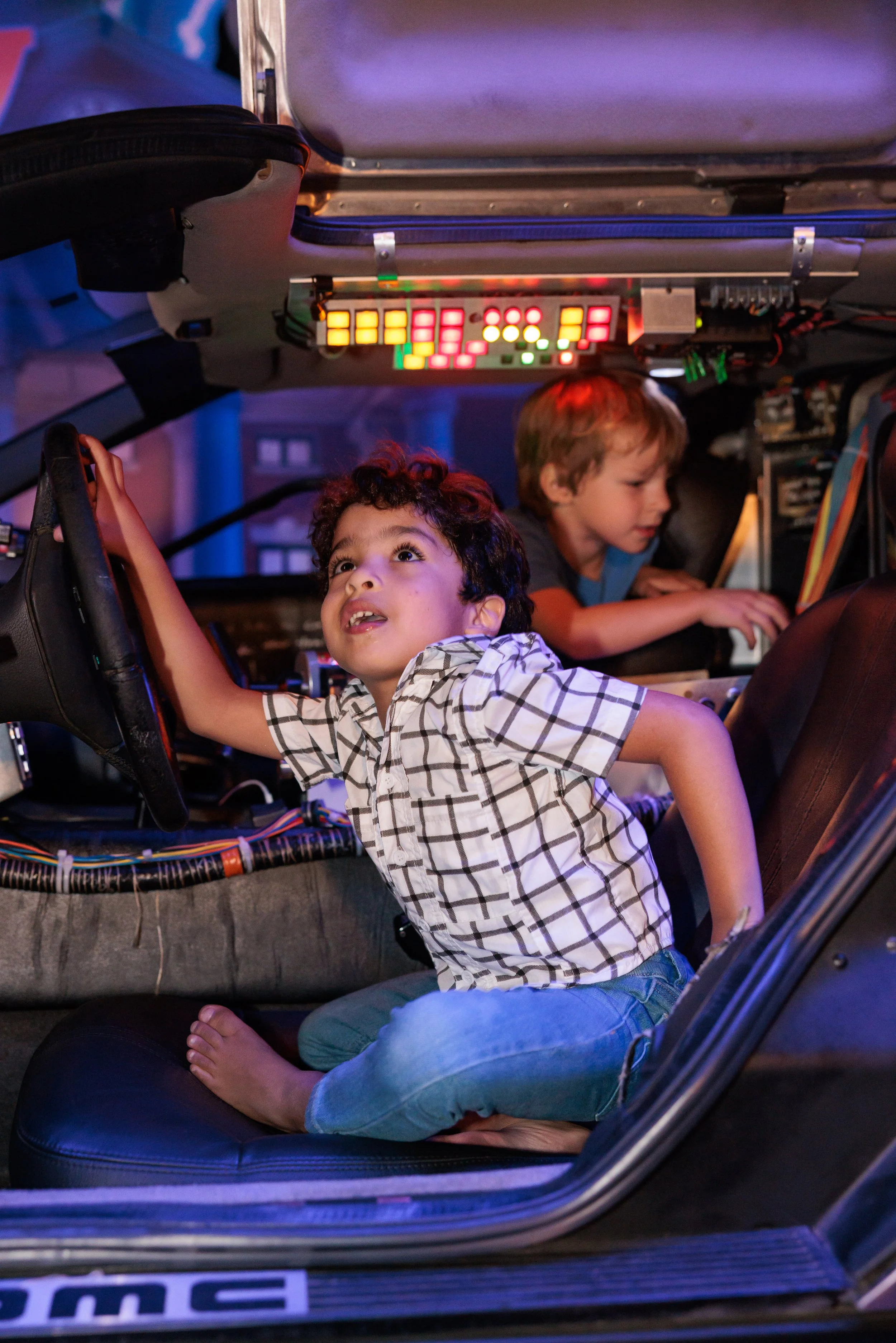 Two young boys playing inside a vehicle with electronic controls and colorful lights, one boy is sitting on the seat with bare feet and reaching for the steering wheel, the other boy is focused on a computer screen.