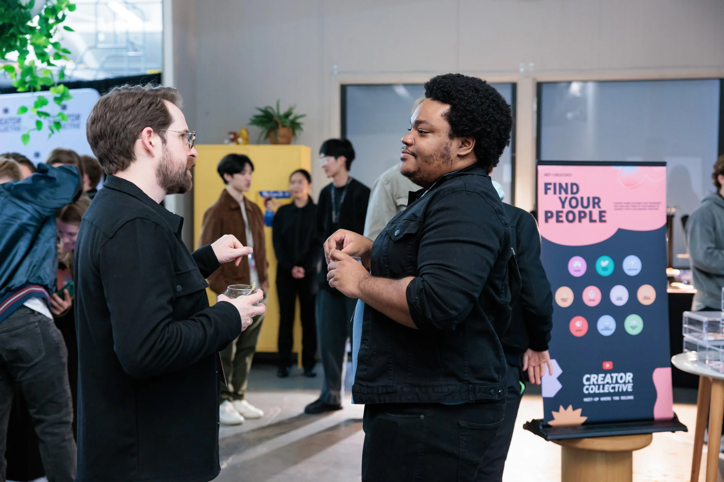 Two men engaged in conversation at a social event, with other attendees in the background and a colorful sign that says 'Find Your People'.