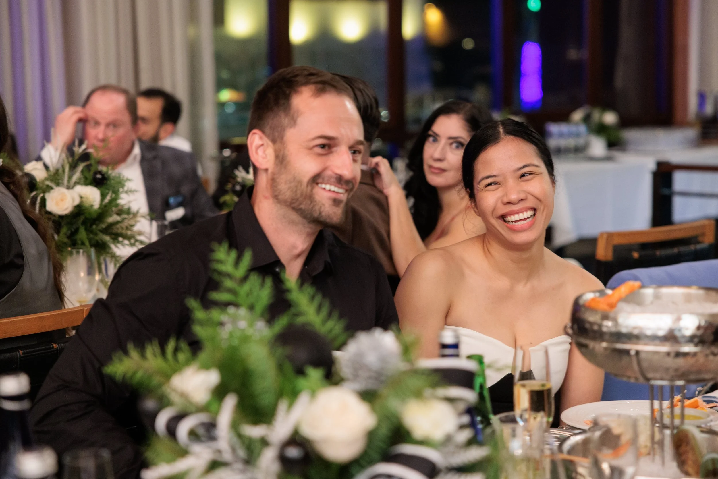 Happy people at a formal dinner event, including a man and a woman in the foreground, smiling and laughing, with other guests and floral centerpieces in the background.