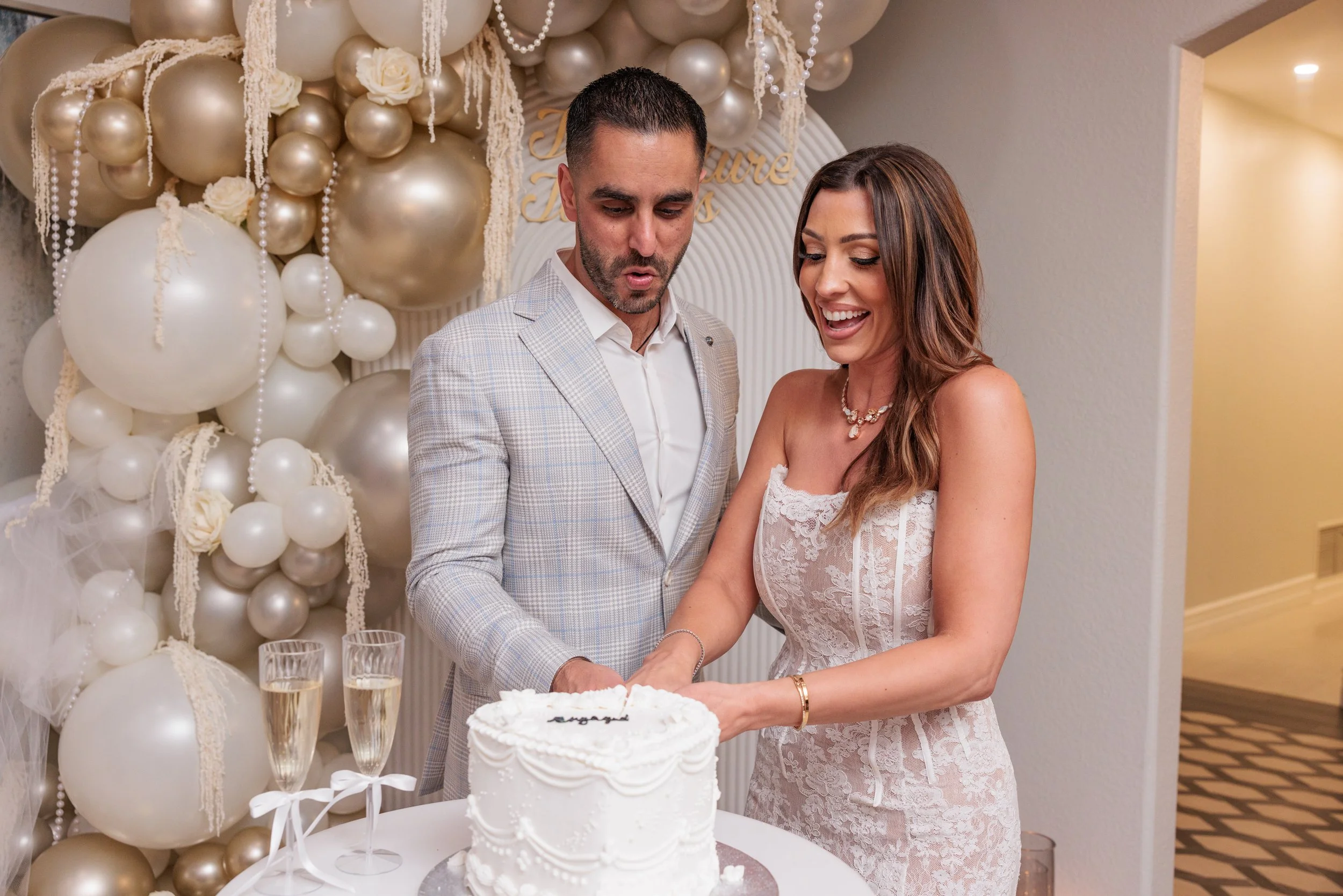 A couple in formal attire cutting a wedding cake at their wedding celebration, with a backdrop decorated with white and gold balloons and flowers.