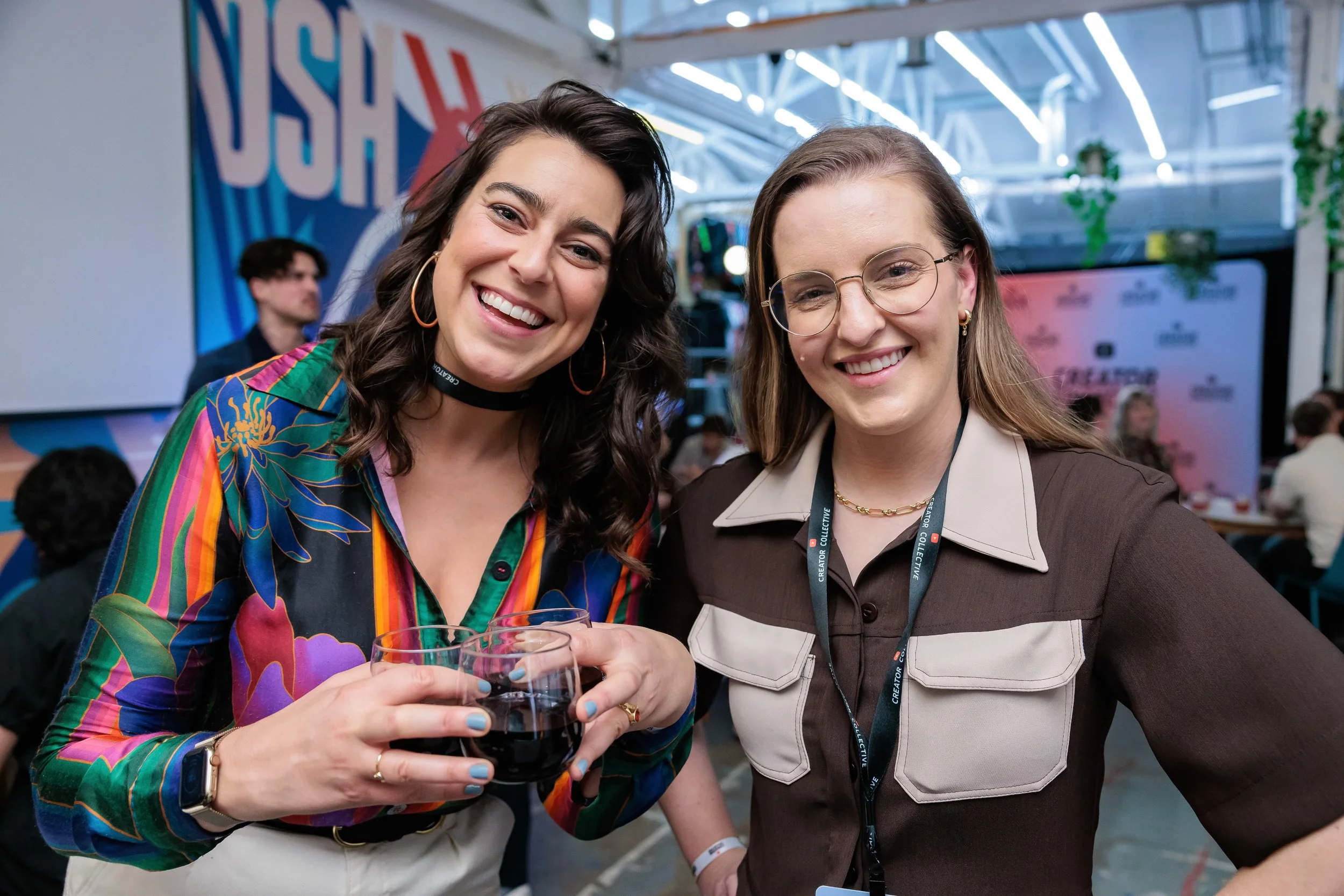 Two women smiling at an event, holding glasses of red wine, with a colorful background and other people in the background.