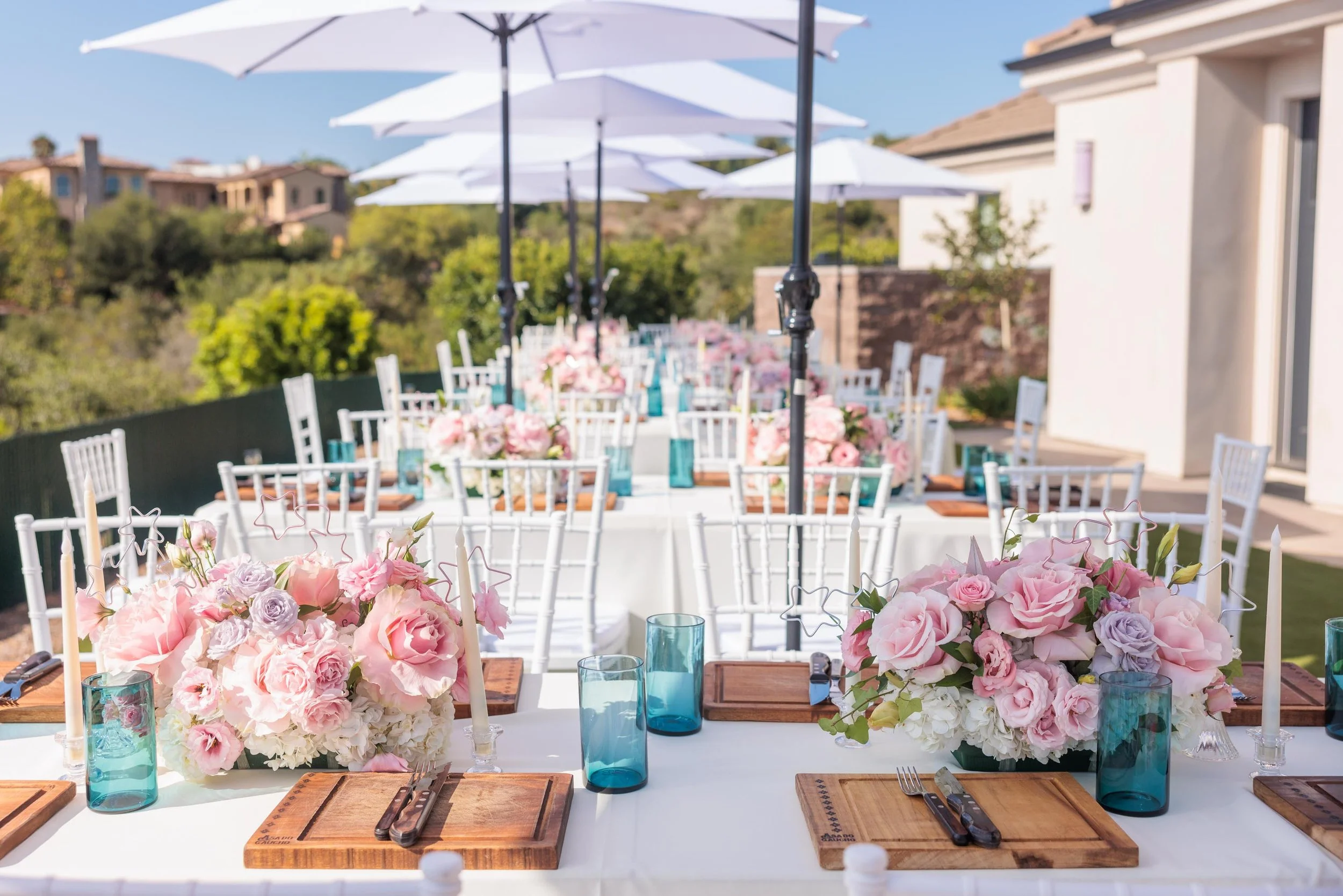 Outdoor wedding reception table decorated with pink and white floral centerpieces, blue glassware, wooden placemats, and white chairs, set under white umbrellas on a sunny day.