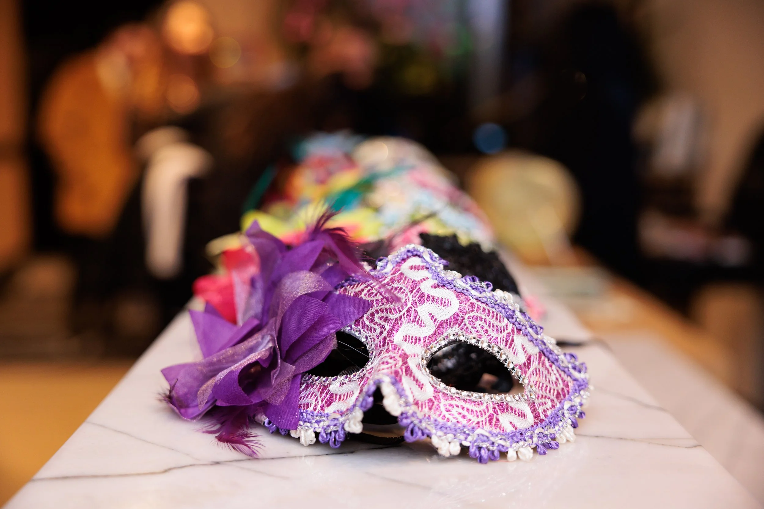 Colorful decorative mask with purple, pink, and white lace, ribbons, and feathers, resting on a white surface.