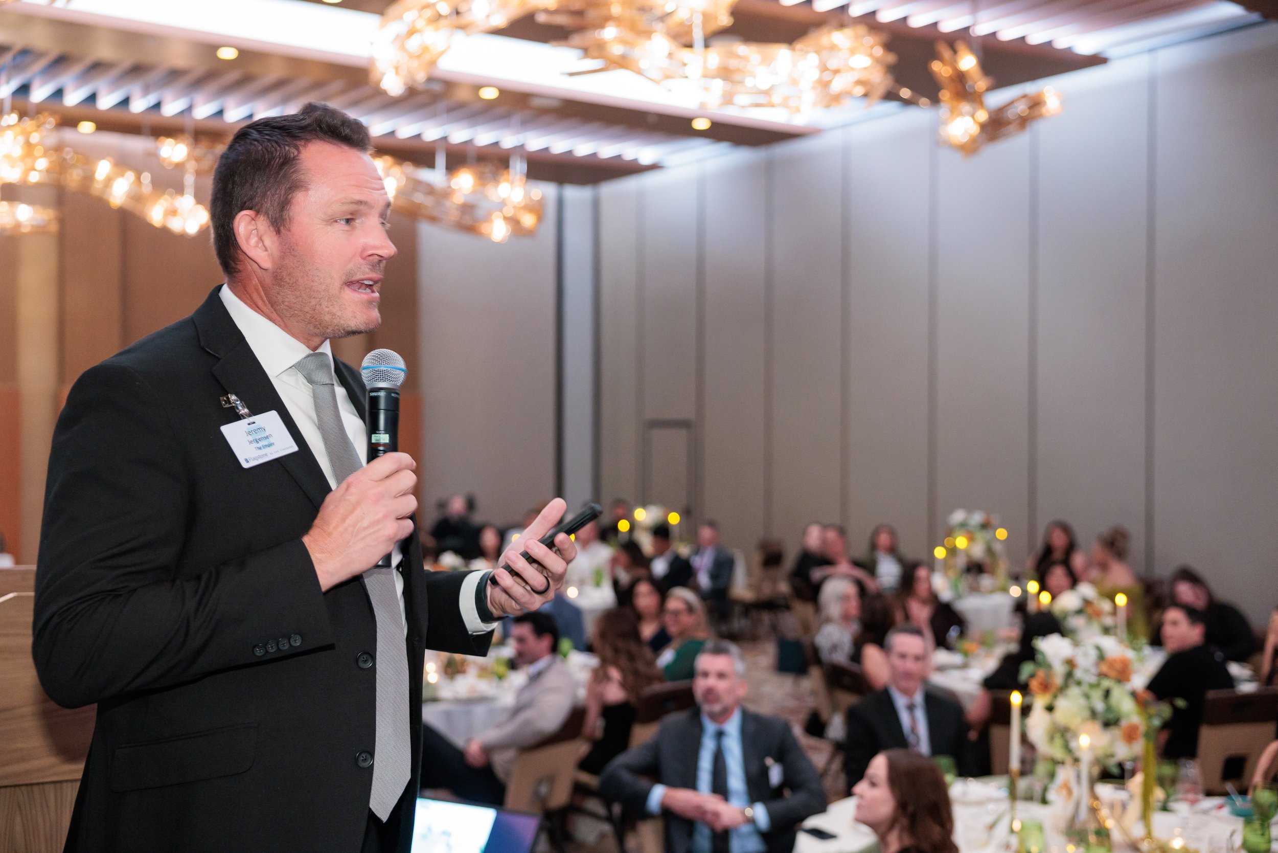 A man in a suit speaking into a microphone at a formal event with many seated guests and floral decorations.