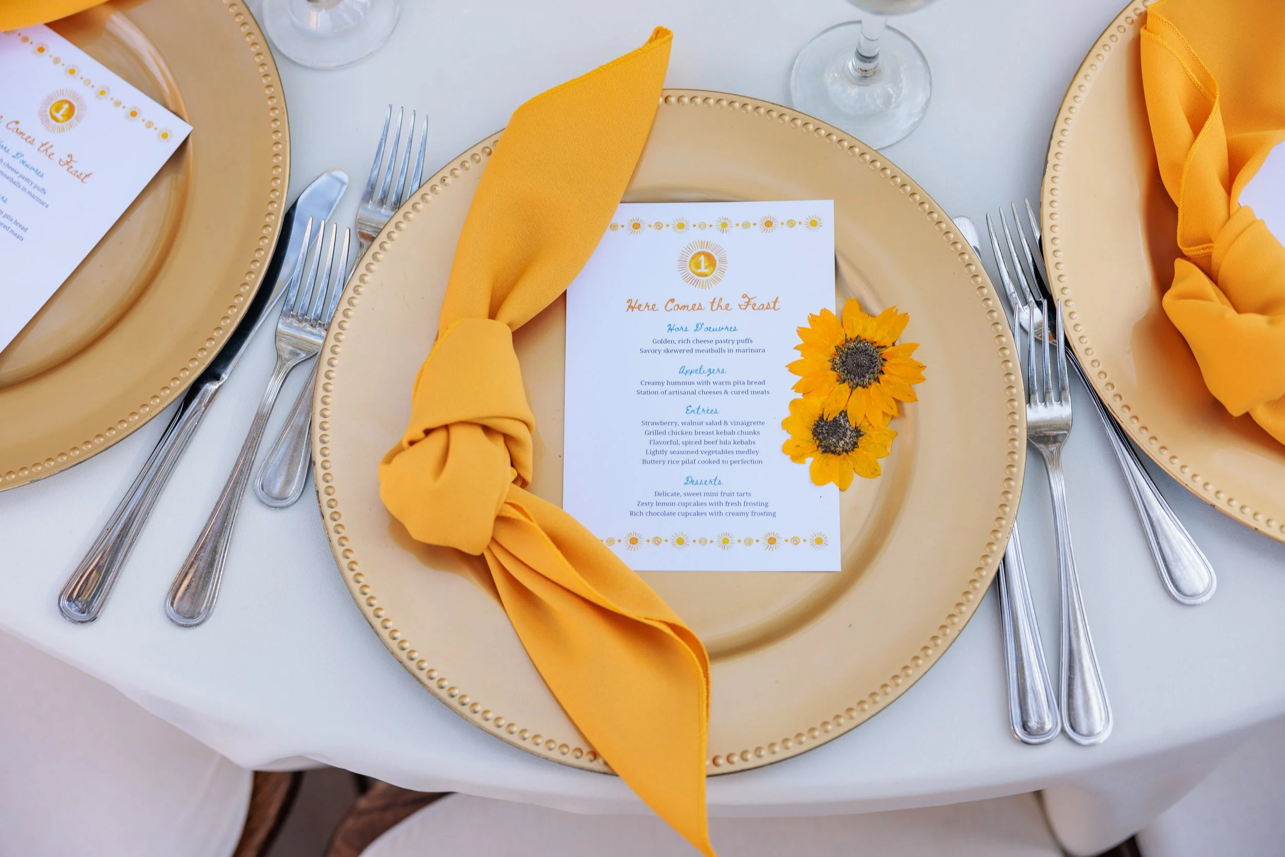 A table setting with a gold charger plate, a white dinner plate, and yellow cloth napkin tied into a knot. There are two forks on the left and a knife on the right of the plates. A menu card with a sunflower decoration is placed on the plate. The tab
