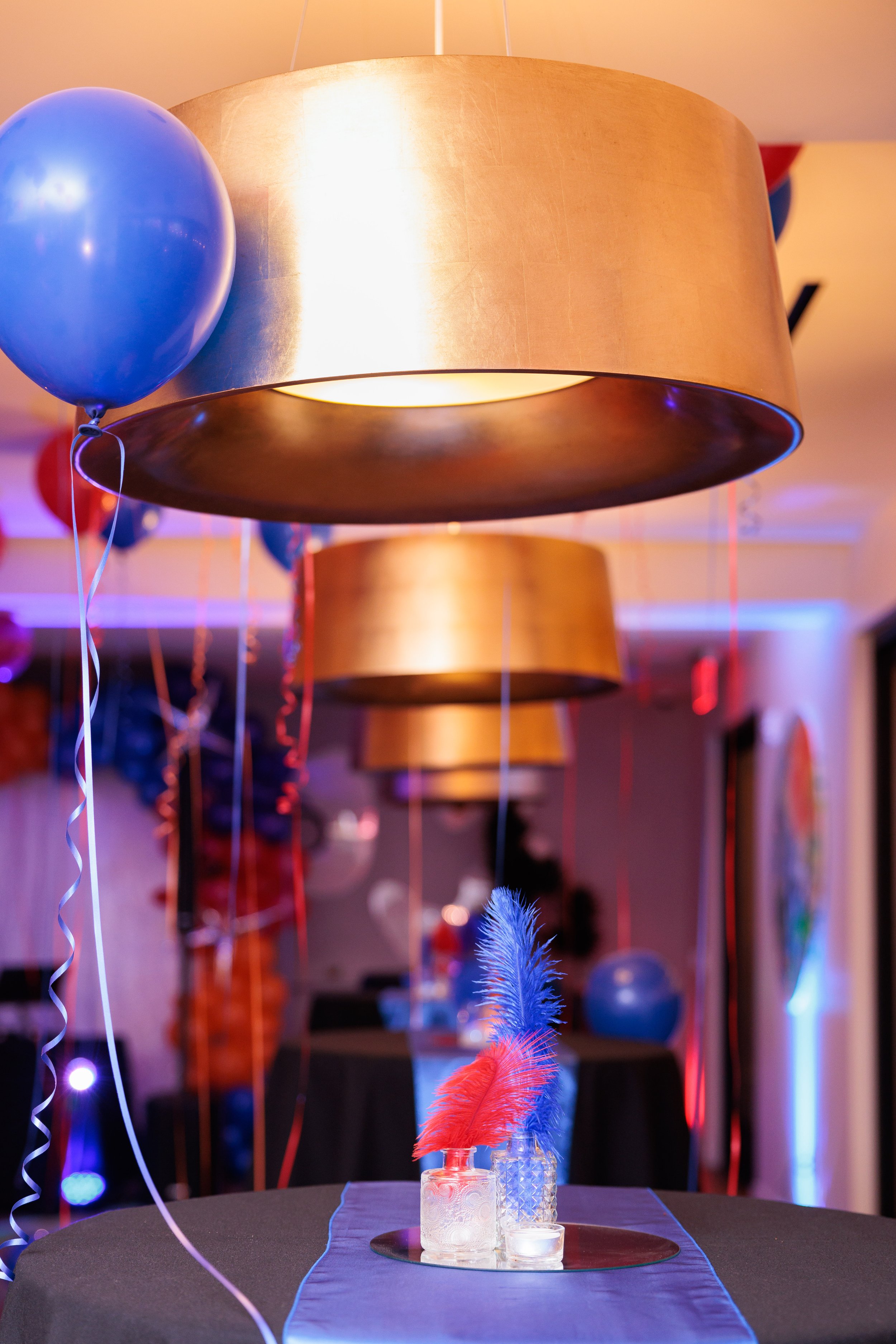Decorative party scene featuring large gold hanging lamps, blue and red balloons, and vases with colorful feathers on a black table with a blue table runner.