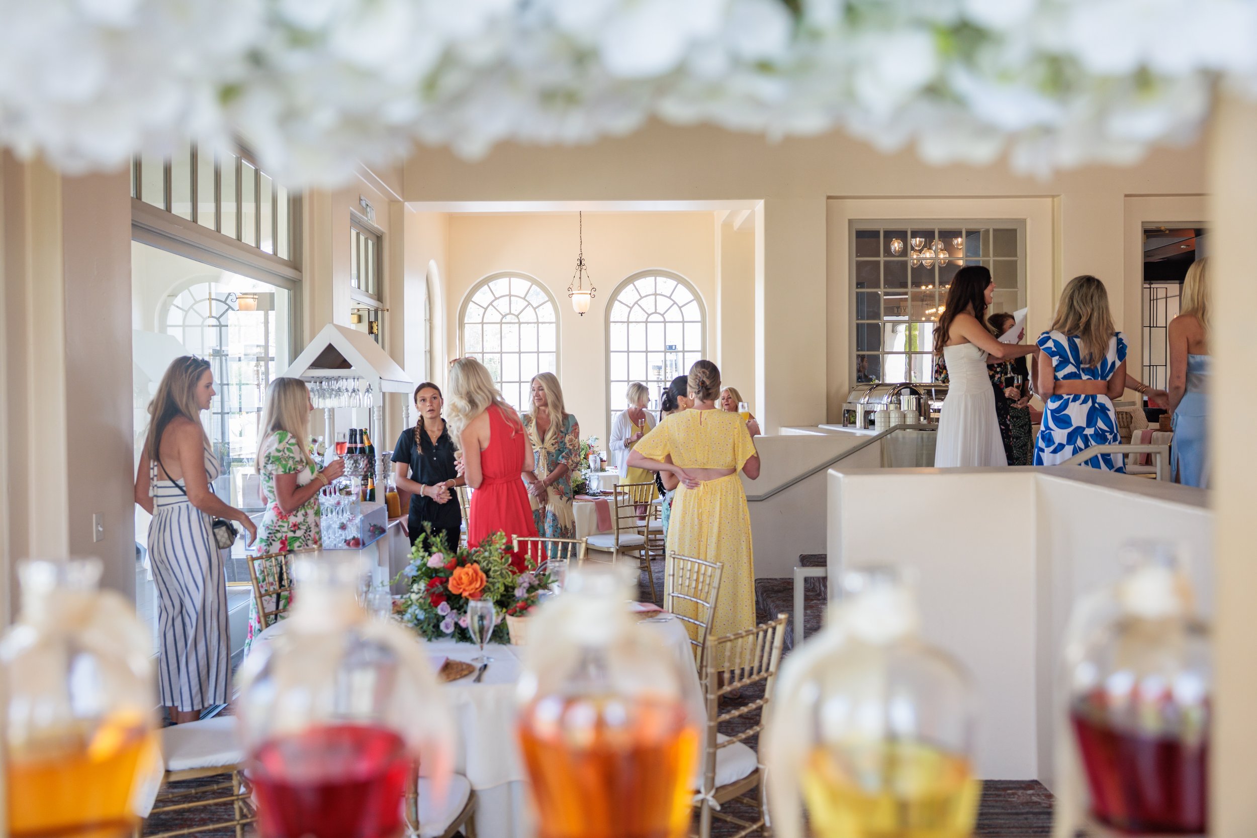 Guests at a daytime social gathering or celebration in a bright, elegant room with large arched windows, some standing around tables decorated with flowers, and colorful drinks in bottles in the foreground.