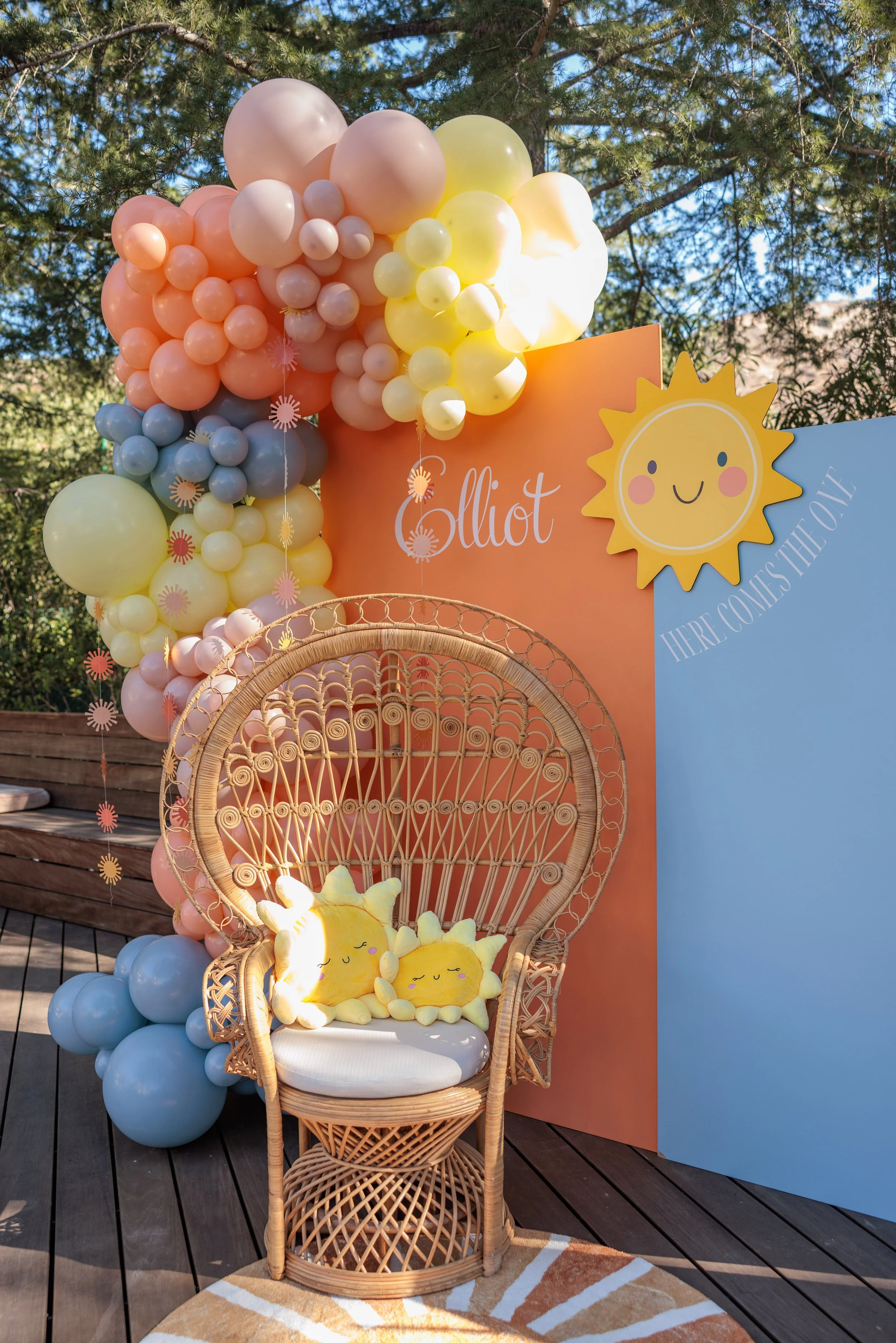 Decorative setup with balloons in pastel colors, a wooden wicker chair with star-shaped plush pillows, and a backdrop with the name 'Elliot' and a smiling sun graphic, likely for a celebration or party.