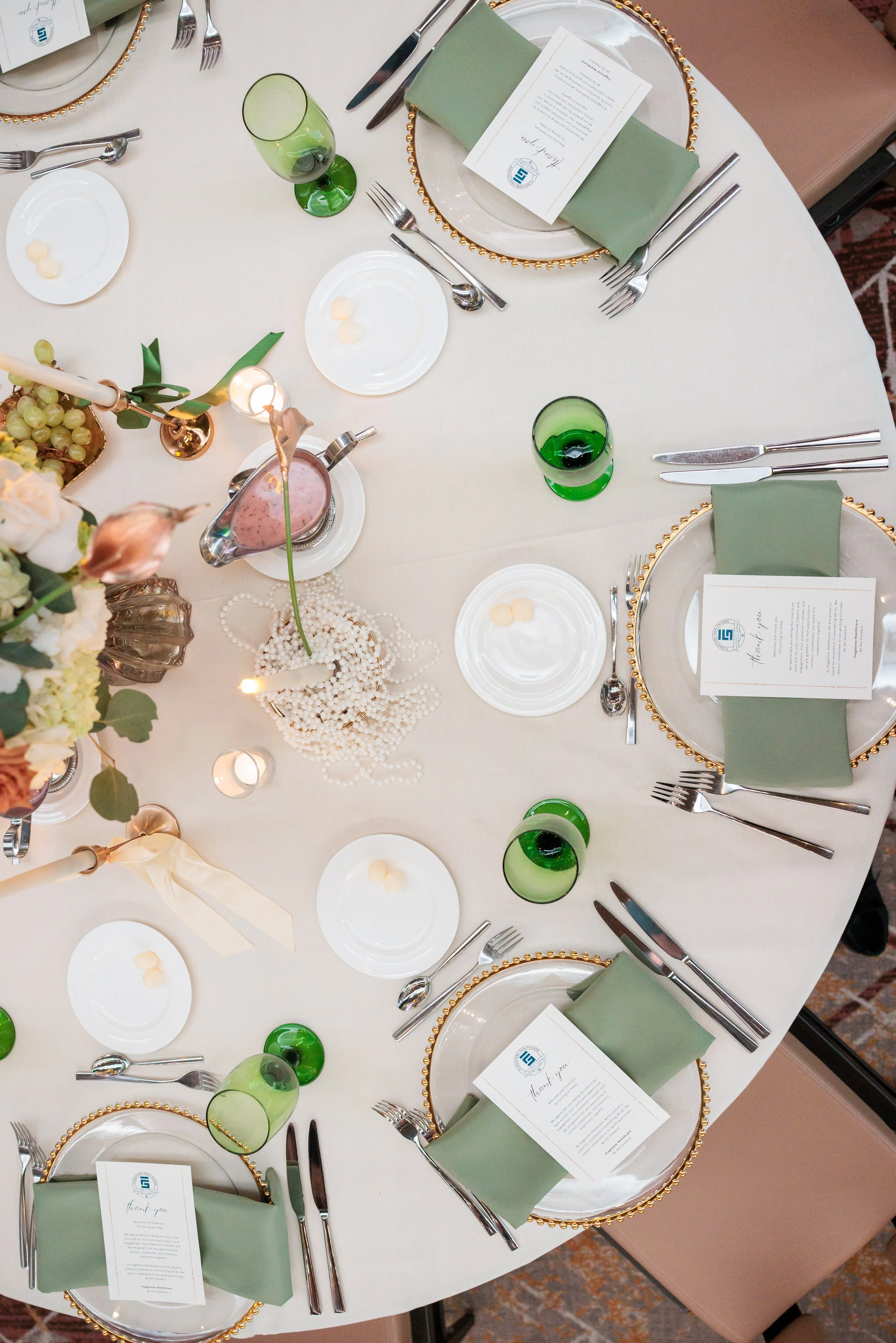 A round banquet table set for a formal event with white tablecloth, green napkins, white dinner plates with gold beaded rims, silverware, green glass tumblers, white plates with butter, floral centerpiece, candles, and thank you cards placed on each 