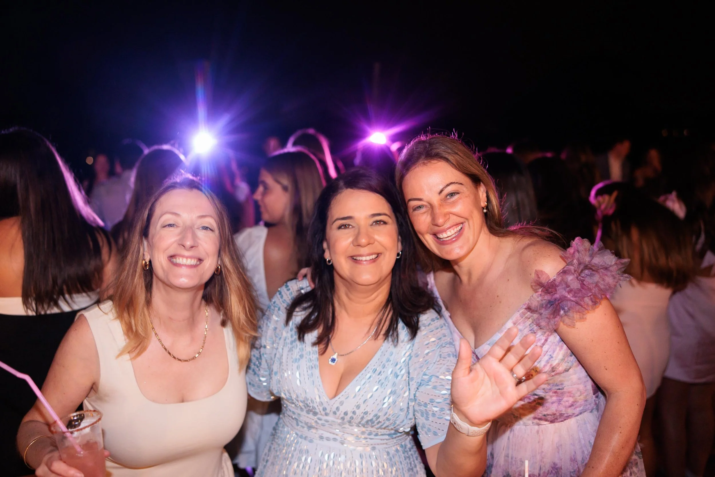Three women smiling and posing together at a nighttime outdoor event or party, with purple and pink lights illuminating the background.