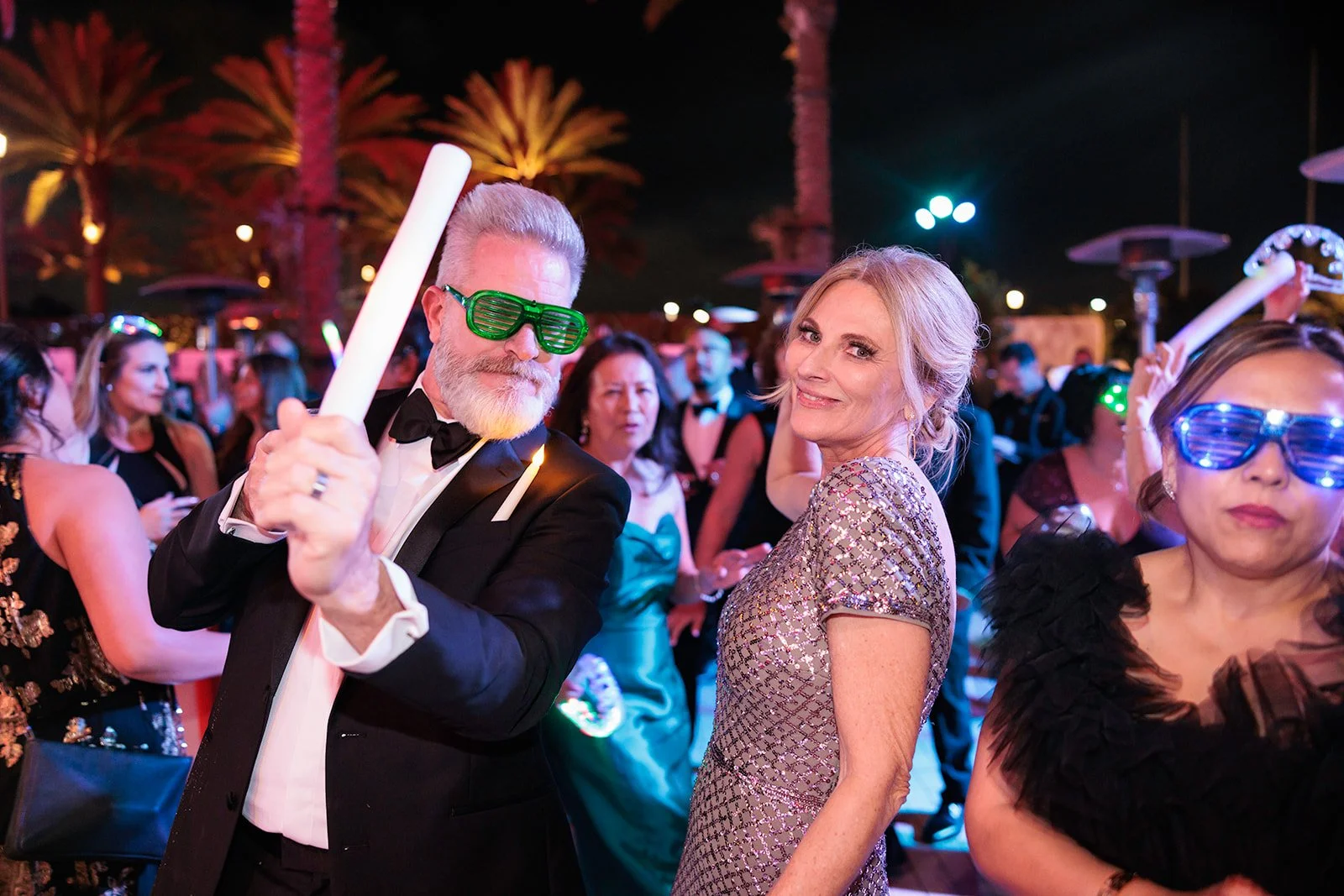People at a fancy party wearing glowing accessories, with some wearing light-up glasses and holding glow sticks, dancing and socializing outdoors at night.
