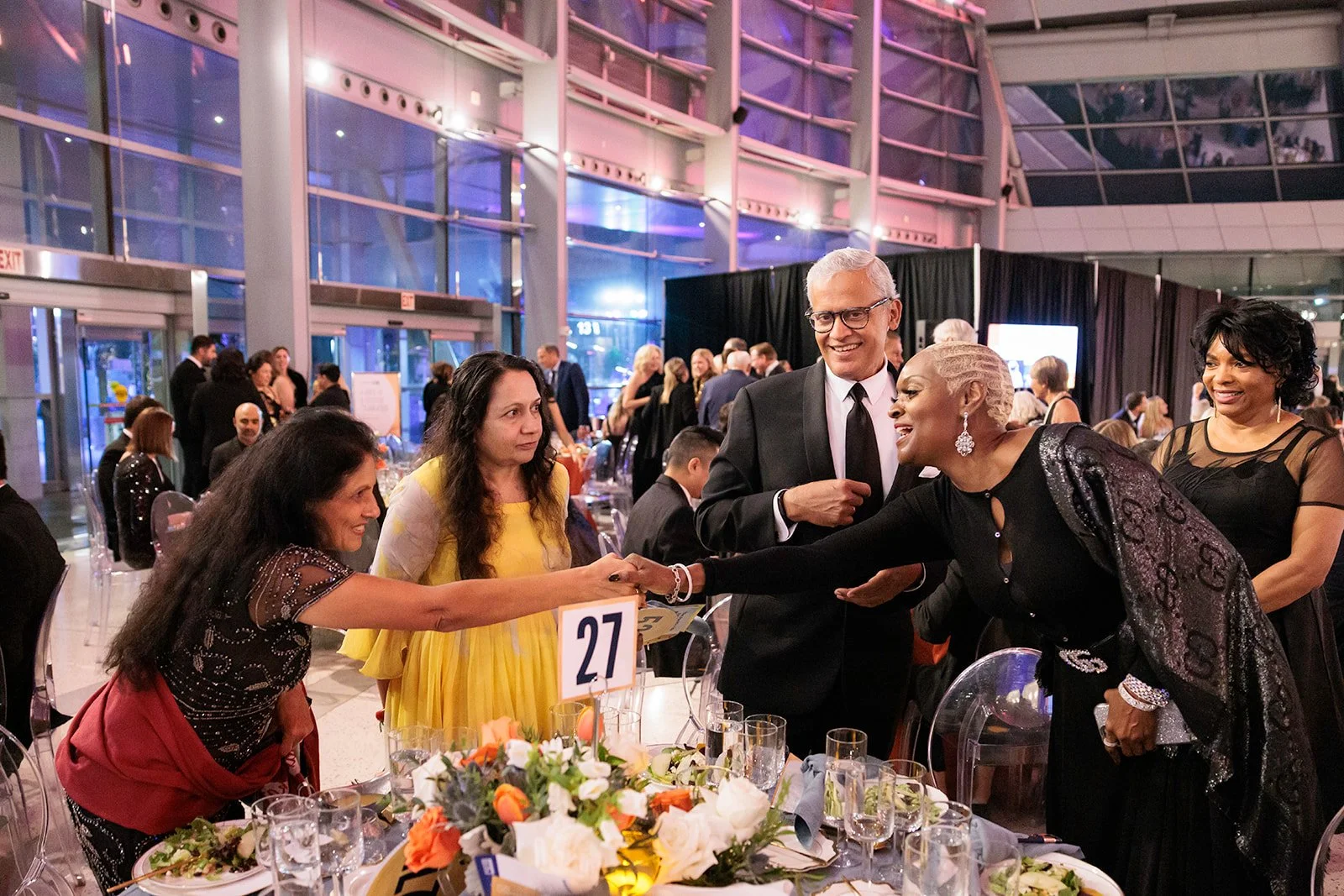 People at a formal event, with a woman in a black dress with glittery sleeves shaking hands with a woman in a yellow dress, surrounded by others talking and smiling at a decorated table in a large glass-walled venue.