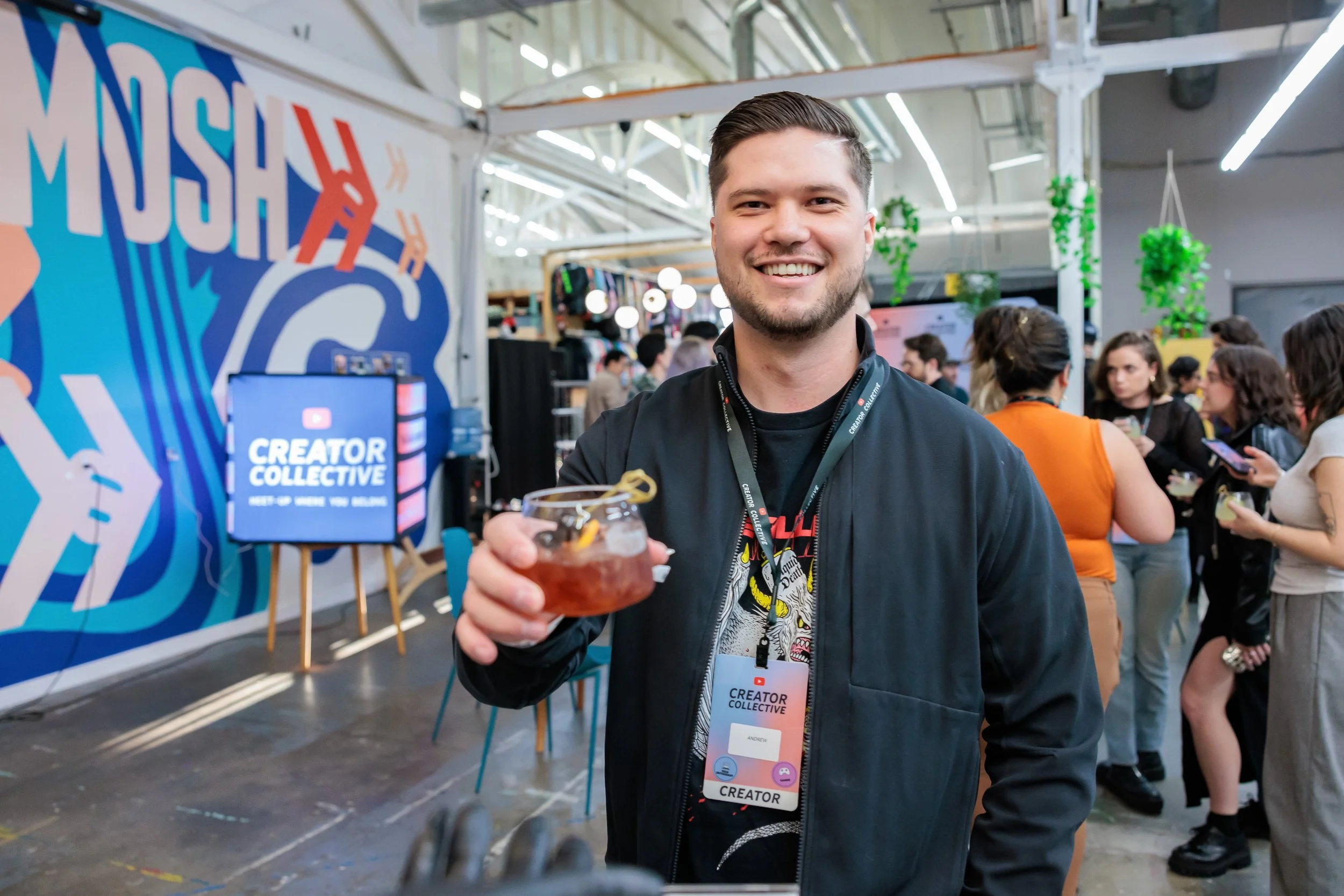 A smiling man holding a cocktail at a creative event called Creator Collective, with a large colorful mural and other people in the background.