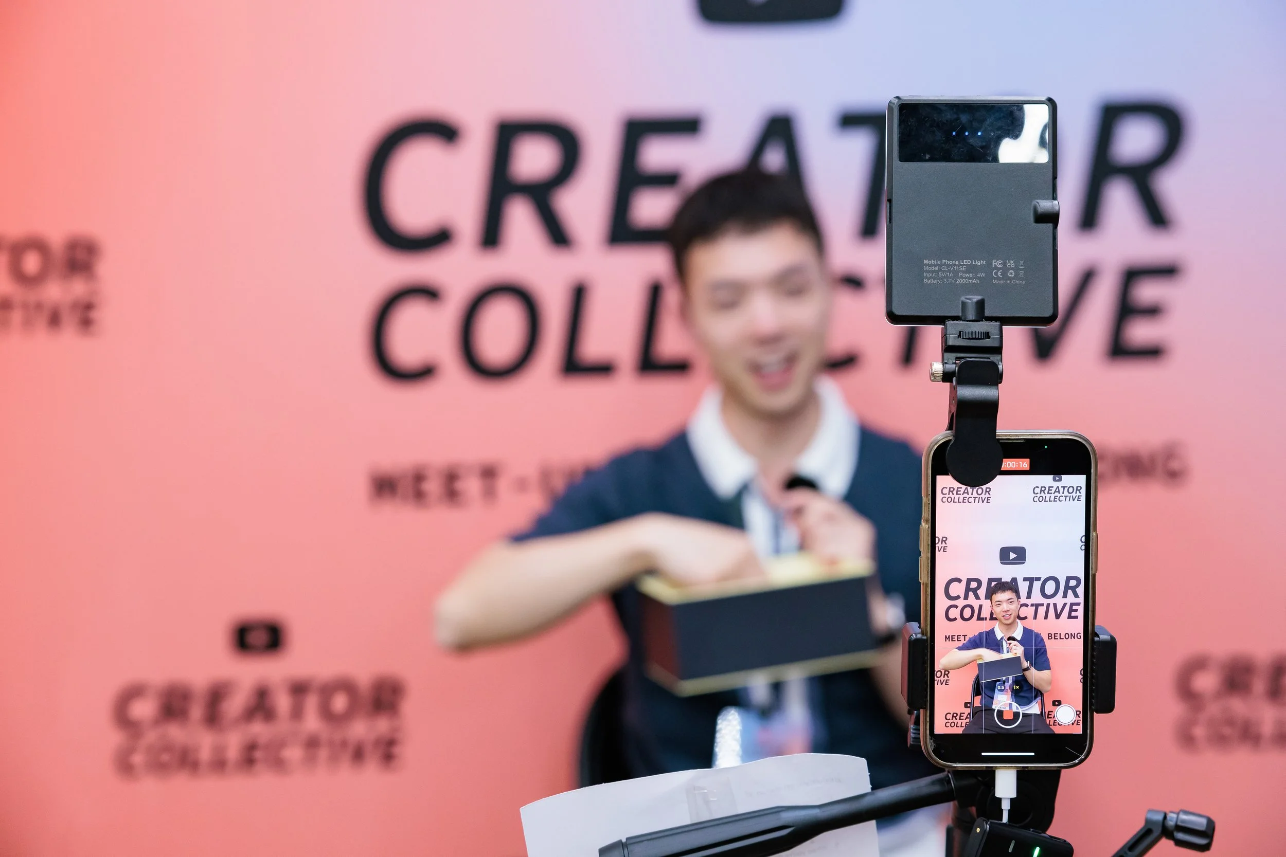 A man recording a video with his smartphone on a tripod, with a pink backdrop that reads 'CREATE, COLLECTIVE' and 'MEET - SHARE - BELONG.'