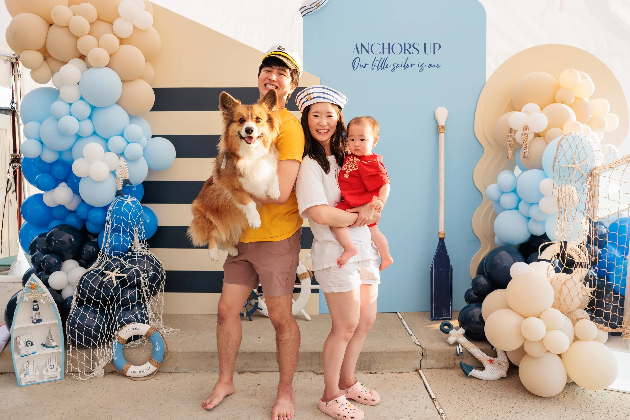 A family of three and a dog at a nautical-themed party with decorations including balloons, a paddle, a life preserver, and a backdrop with the text 'Anchors Up, Our little sailor is one'.