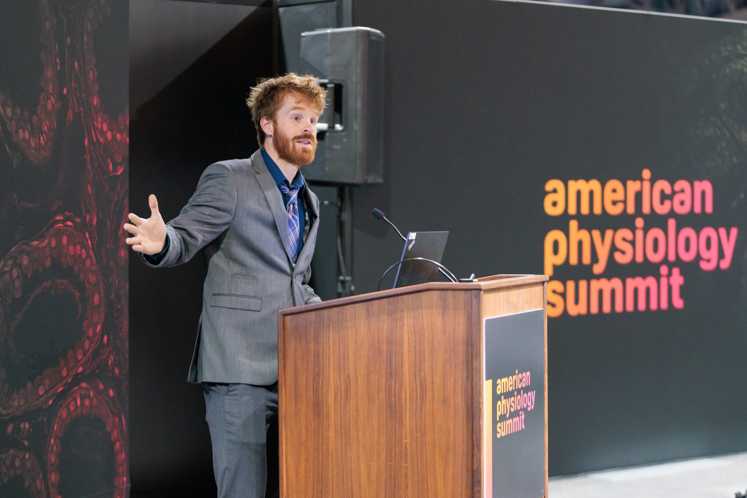 A man in a gray suit giving a presentation at the American Physiology Summit, standing behind a wooden podium with a laptop, gesturing with one hand.