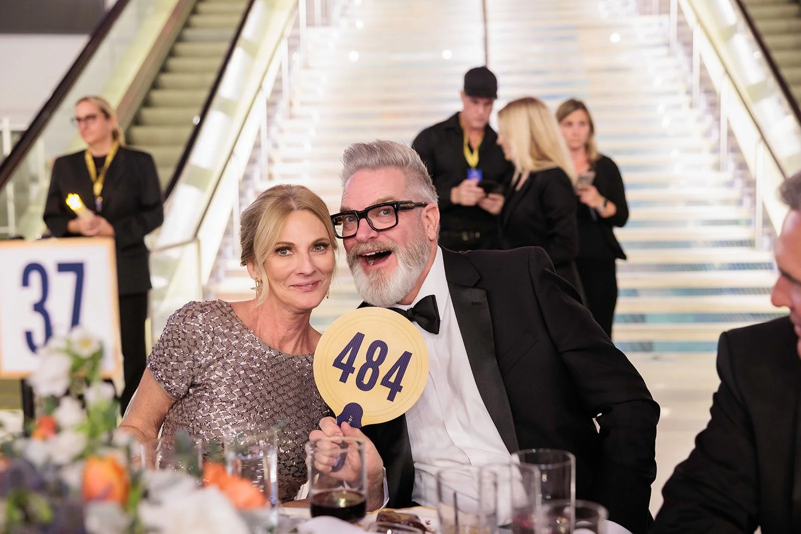 A smiling elderly woman in a patterned dress and a smiling man with glasses, gray hair, and a beard wearing a tuxedo, holding a paddle with the number 484 at a formal event. In the background, other guests are seated at a table near a staircase.