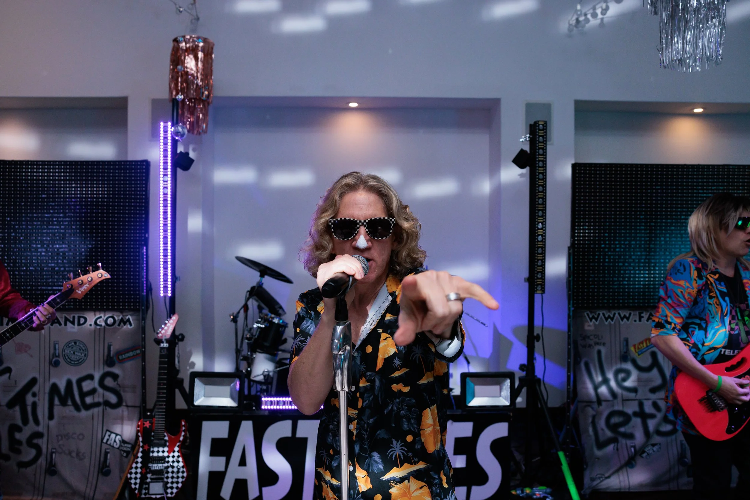 A woman wearing sunglasses and a Hawaiian shirt sings into a microphone on stage during a band performance, with band members and musical equipment visible in the background.