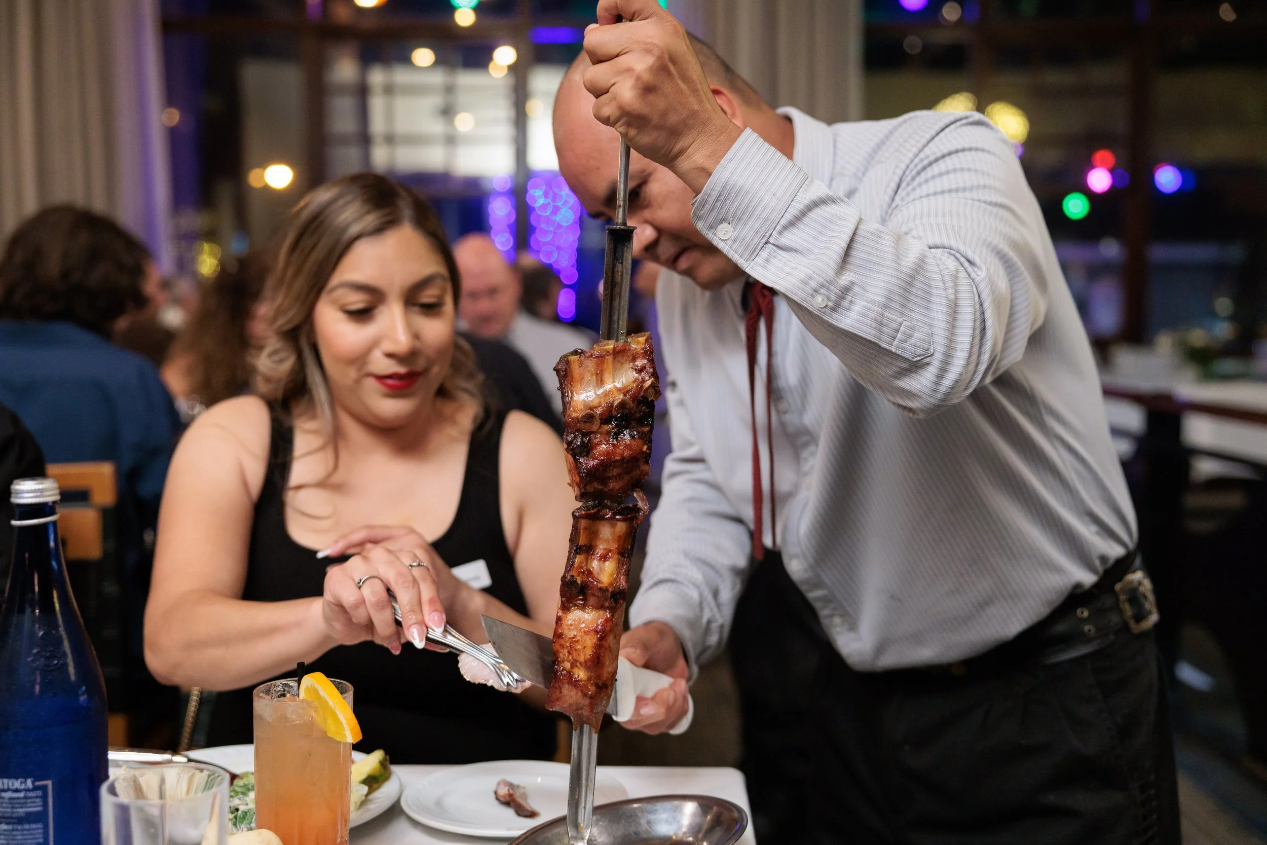 A man serving ribs on a skewer at a restaurant while a woman watches and prepares to cut a piece, with other diners in the background.