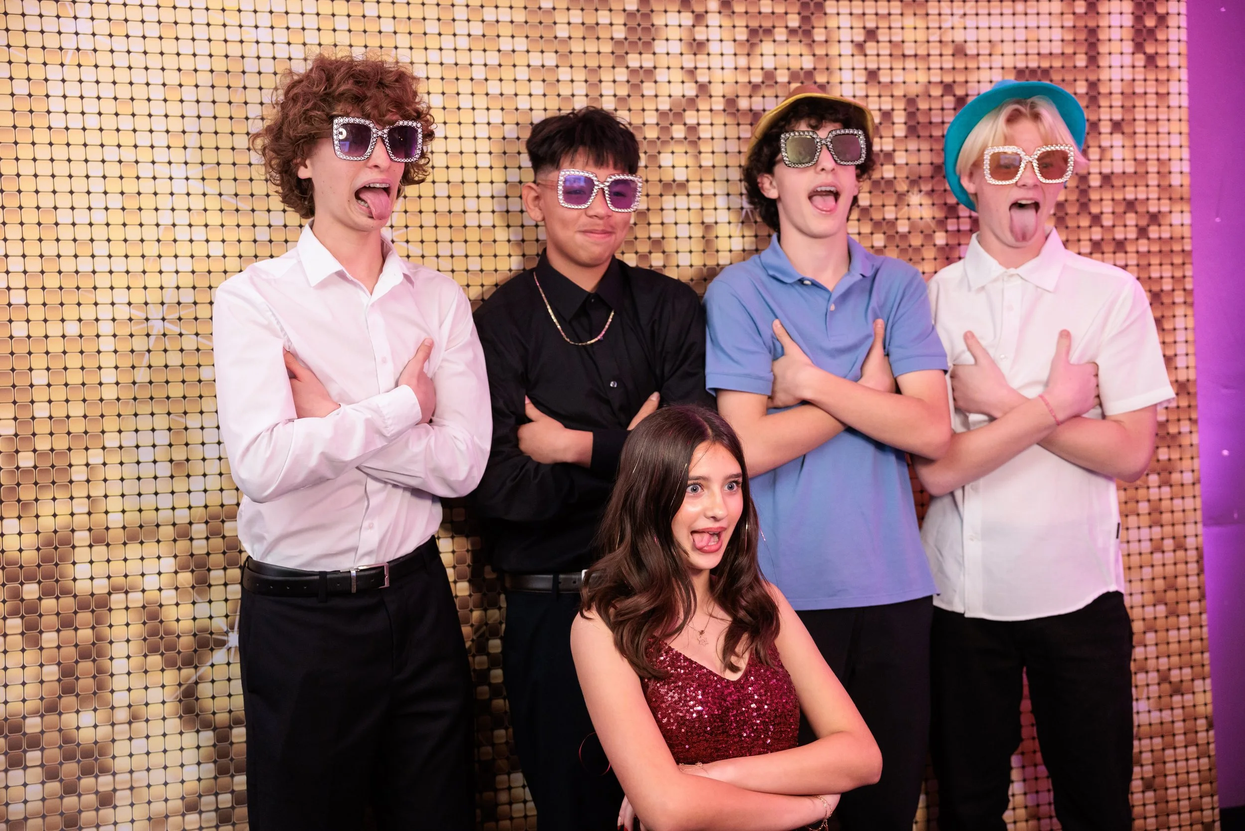 Group of five teenagers posing funnily with sunglasses in front of a sparkly gold backdrop.
