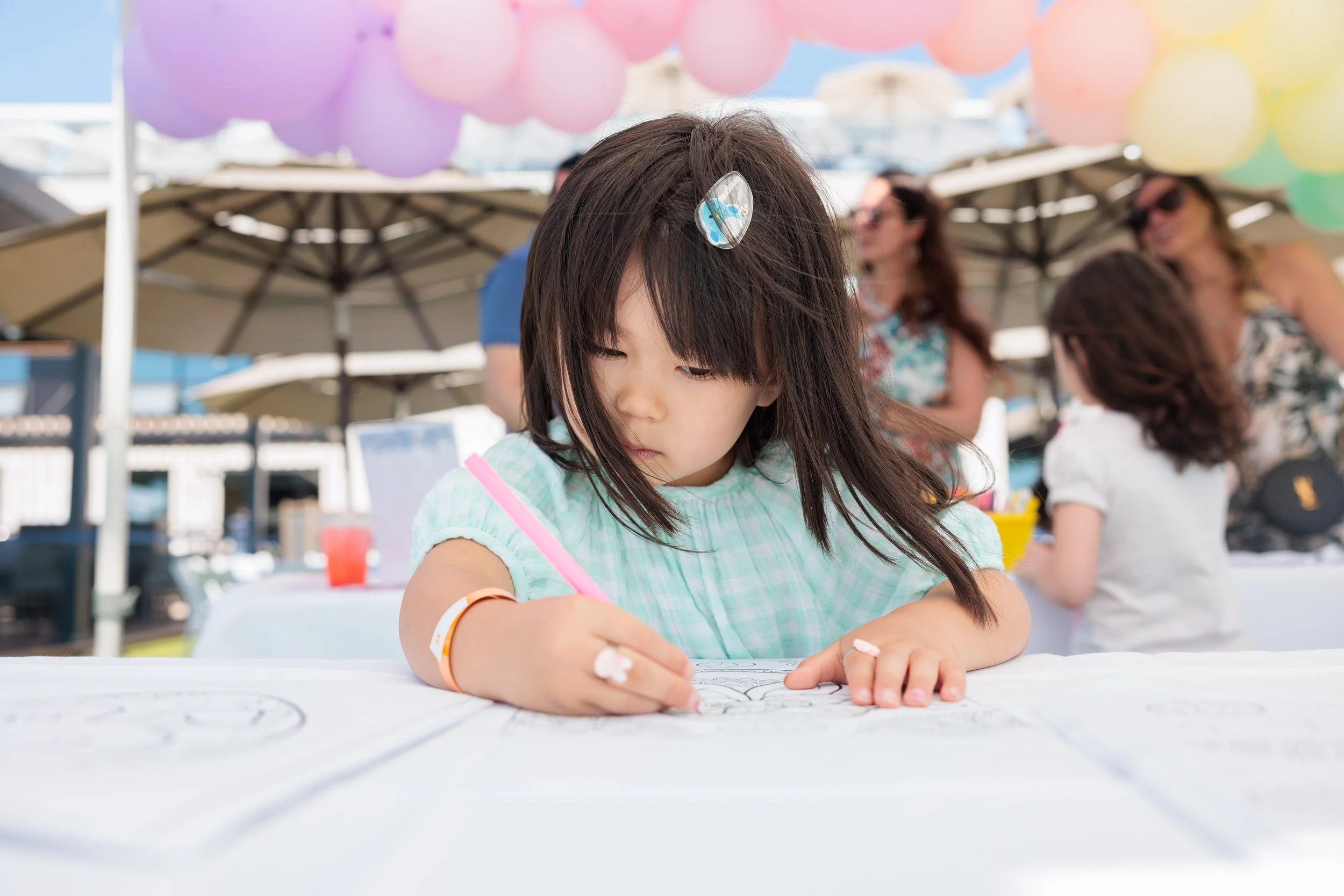 A young girl with a small pink marker drawing or coloring on a large white sheet of paper at an outdoor event, with people and colorful balloons in the background.
