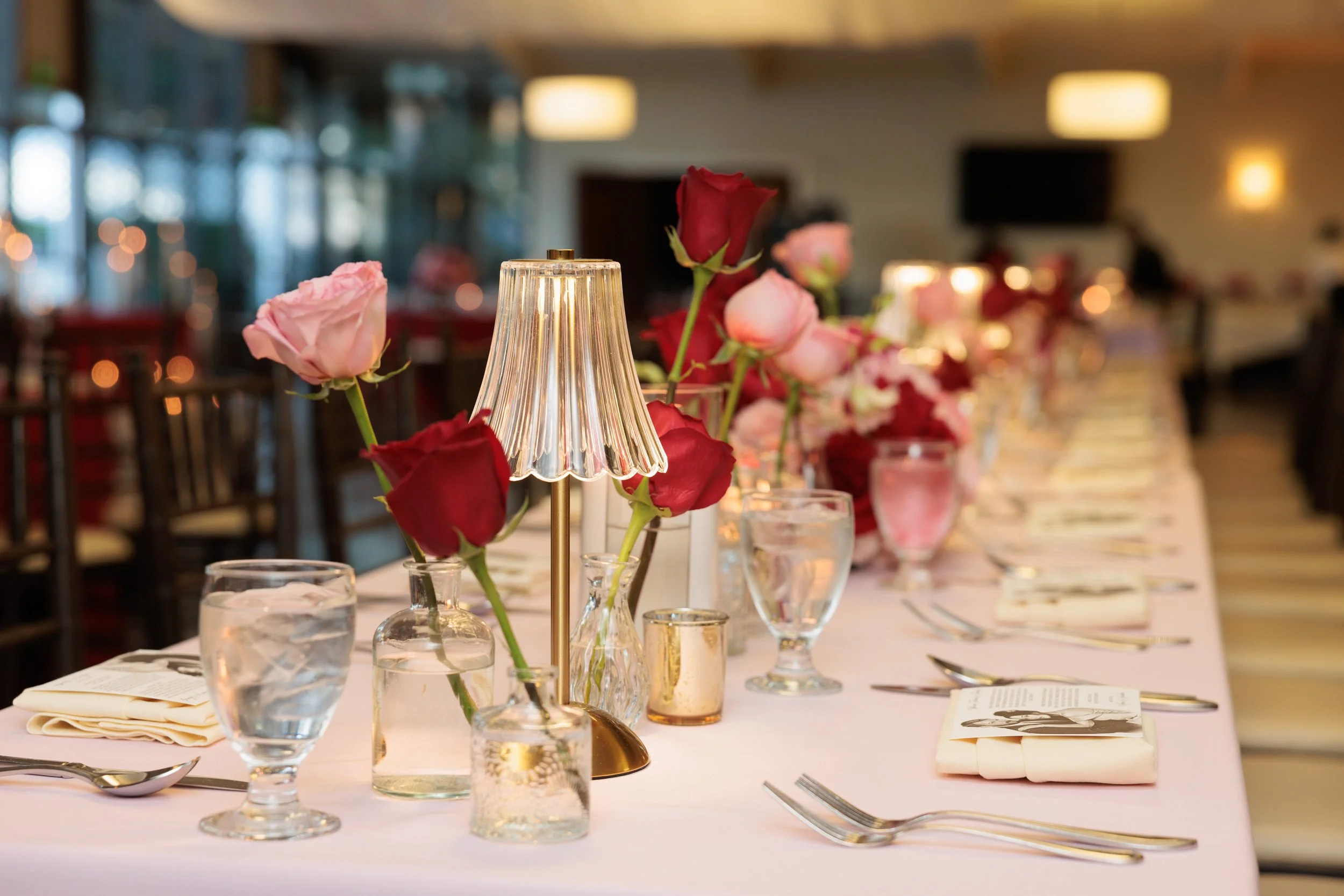 A long dining table decorated with pink and red roses in vases, illuminated by a small lamp, set with glassware, cutlery, and folded napkins, in a well-lit banquet hall.