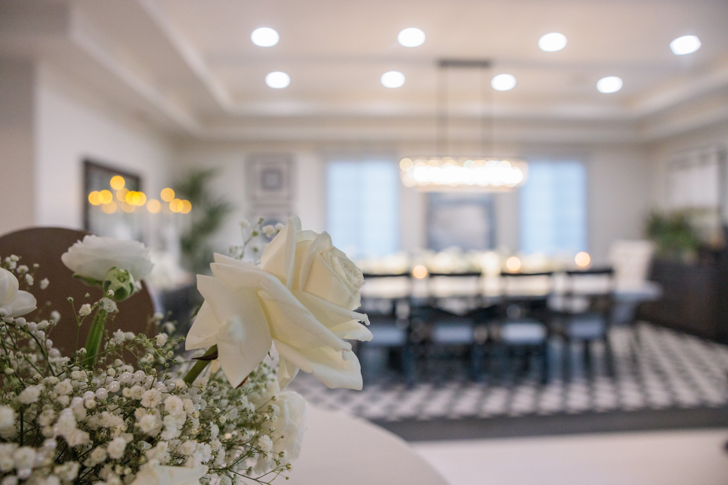 Close-up of white roses and baby's breath flowers on a table with a blurred background of a decorated dining room with a chandelier, windows, and a checkered floor.