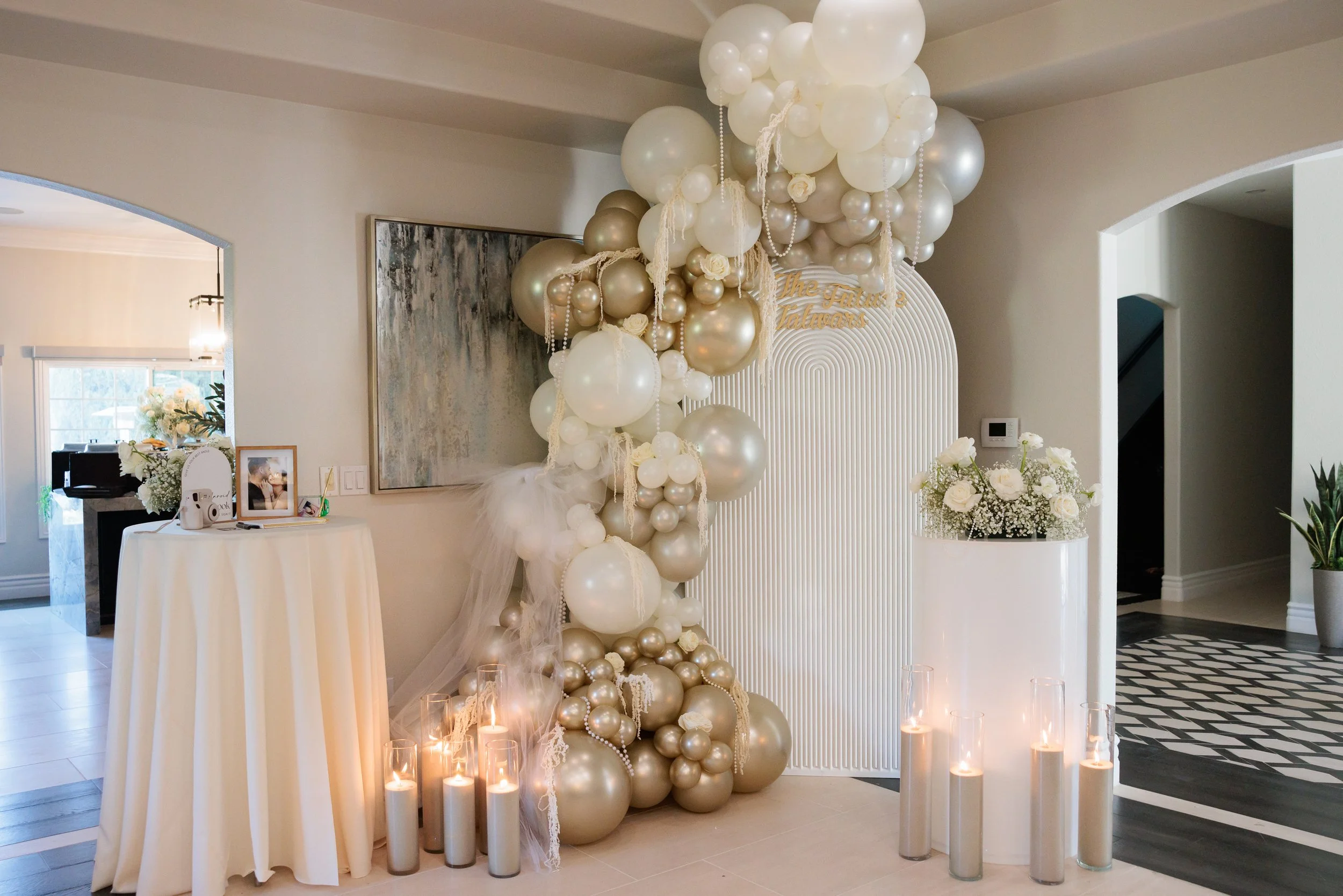 Elegant indoor decoration with white and gold balloons forming a front backdrop, surrounded by tall candles with lit candles and white flowers, for a celebration event.