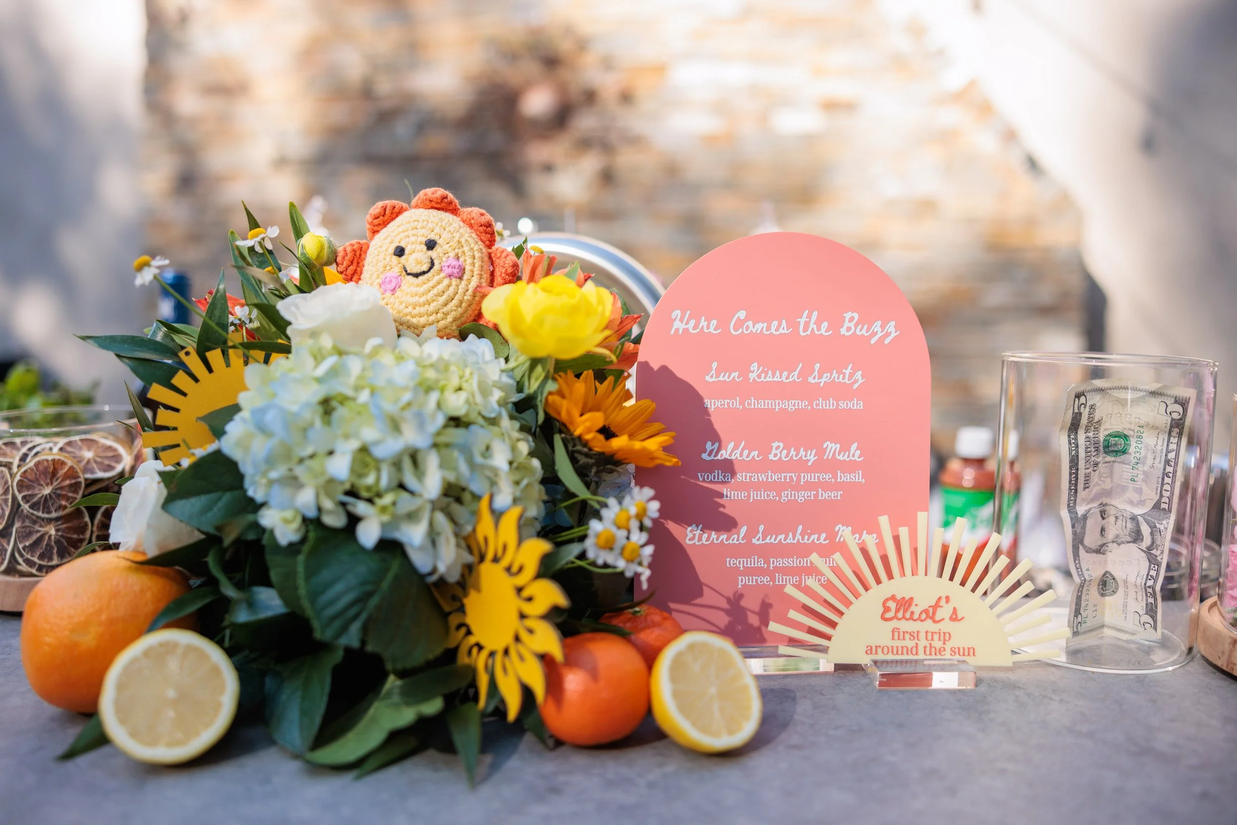 A colorful flower arrangement with a crochet lion toy, citrus fruits, and a pink menu card with drink names. A plastic cup with dollar bills and a small bottle of soda are nearby.