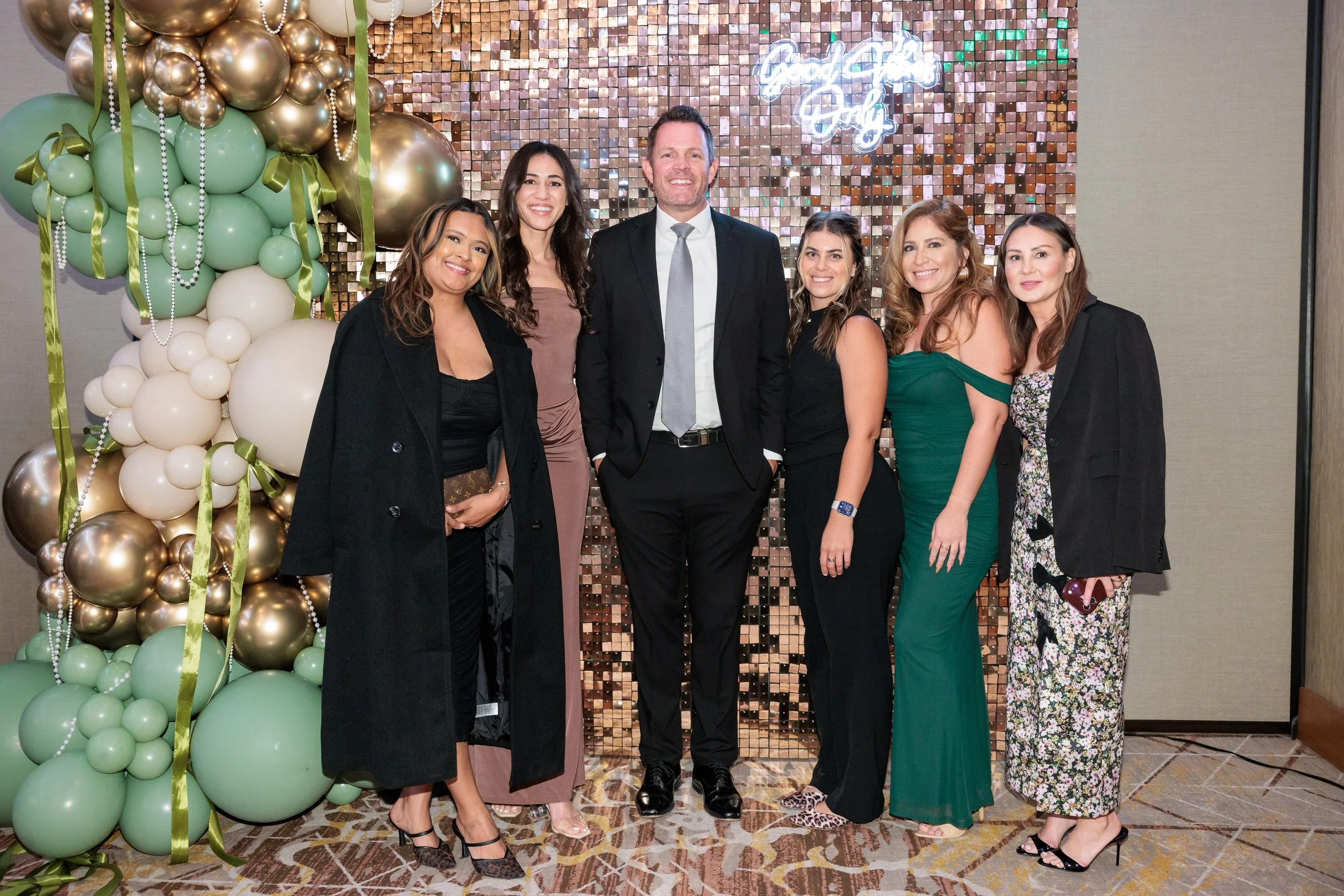 A group of seven people standing in front of a decorative balloon backdrop at a celebration, with a neon sign that reads "Good Vibes Only." They are dressed in formal attire and smiling for the camera.