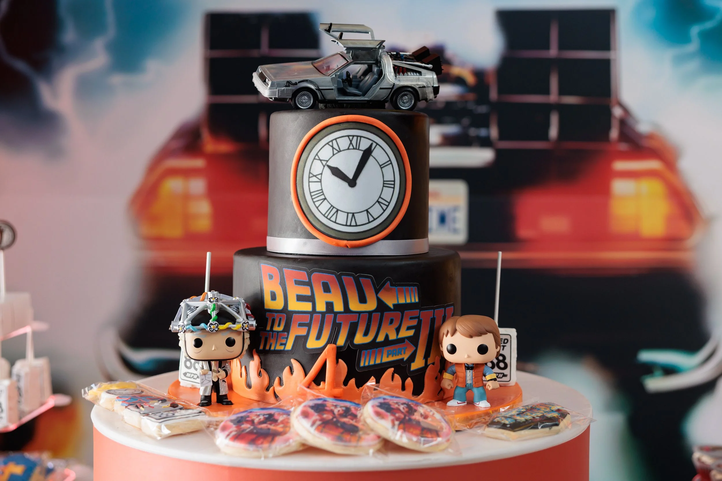 A themed cake for the movie 'Back to the Future' with figurines of Marty McFly and Doc Brown, a DeLorean car model on top, a clock, and assorted cookies.