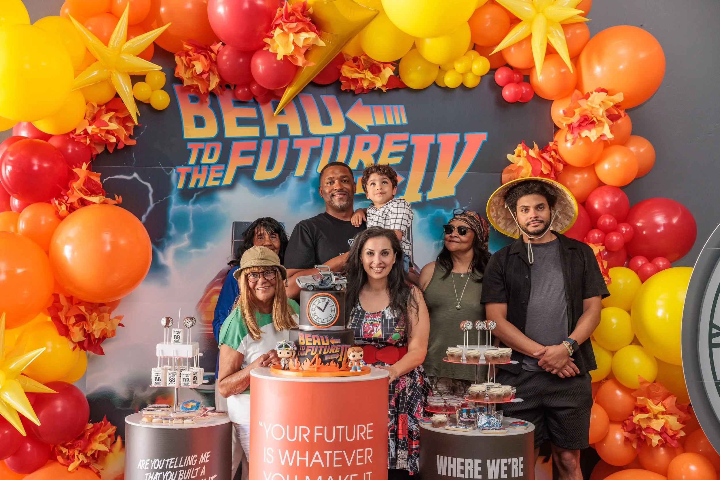 Group of people celebrating a "Back to the Future" themed event with balloons, decorations, and themed treats at a party.