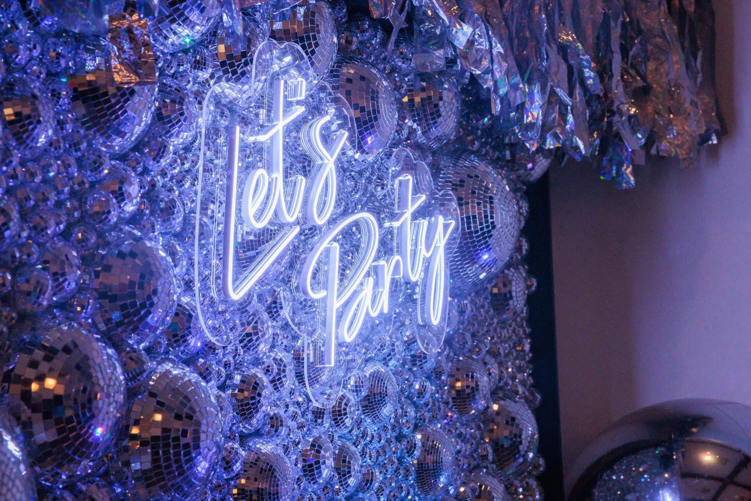 Neon sign reading 'Let's Party' on a background of silver disco balls and metallic decorations.