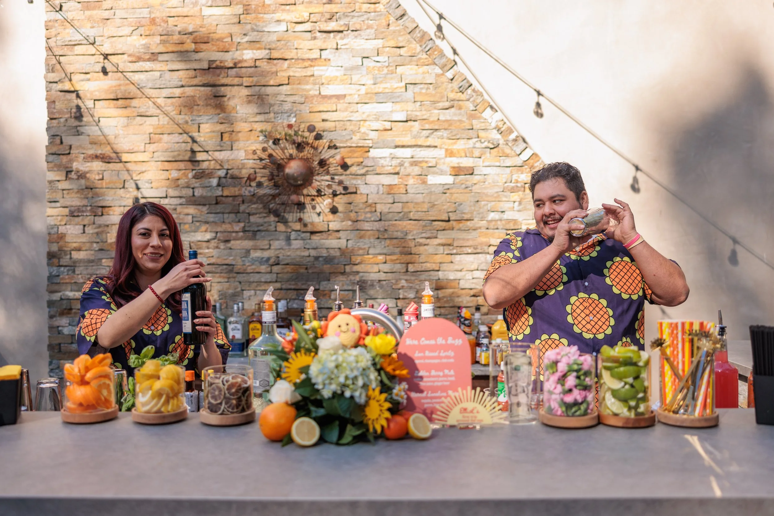 Two bartenders behind a bar, one woman and one man, preparing drinks, with colorful fruit garnishes, bottles of alcohol, and cocktail ingredients on the counter. The woman has long dark hair and the man is smiling, both wearing matching Hawaiian shir