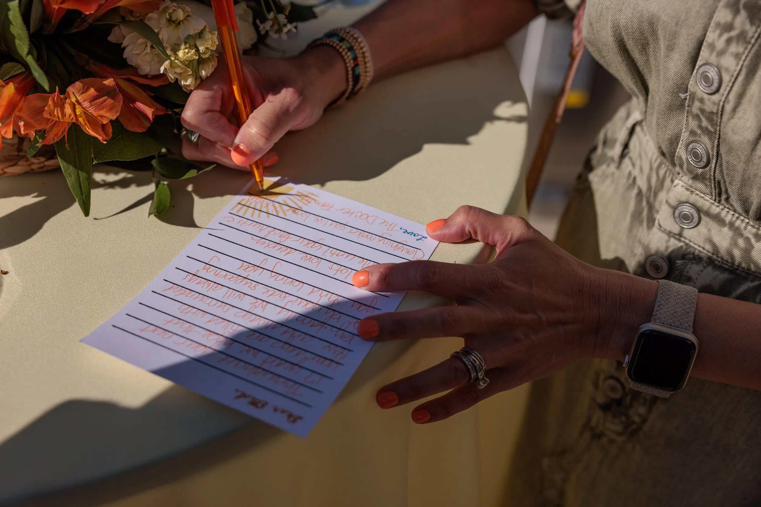 A person writing on a lined white card with orange ink, surrounded by flowers and using an orange pen. The person has orange nails, a ring, a wristwatch, and bracelets. They are wearing a beige buttoned garment.