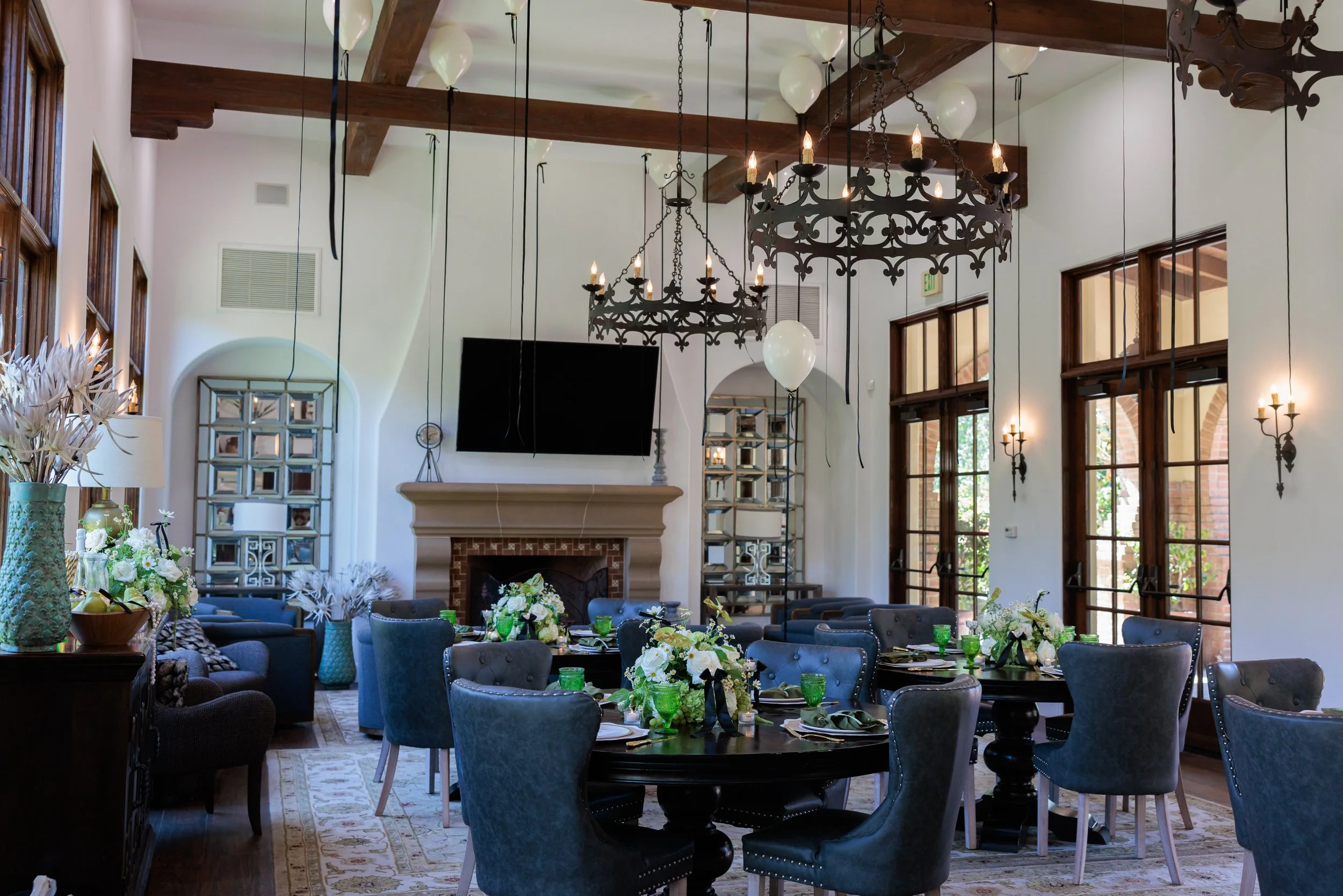 Elegant dining room with round tables, blue chairs, floral centerpieces, large windows, chandelier, balloons, and a fireplace with a TV above it.