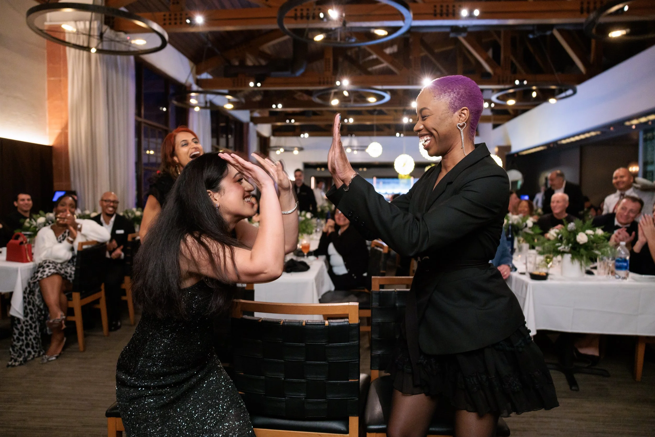Two women celebrating and dancing together at a formal event with other seated guests clapping in the background.