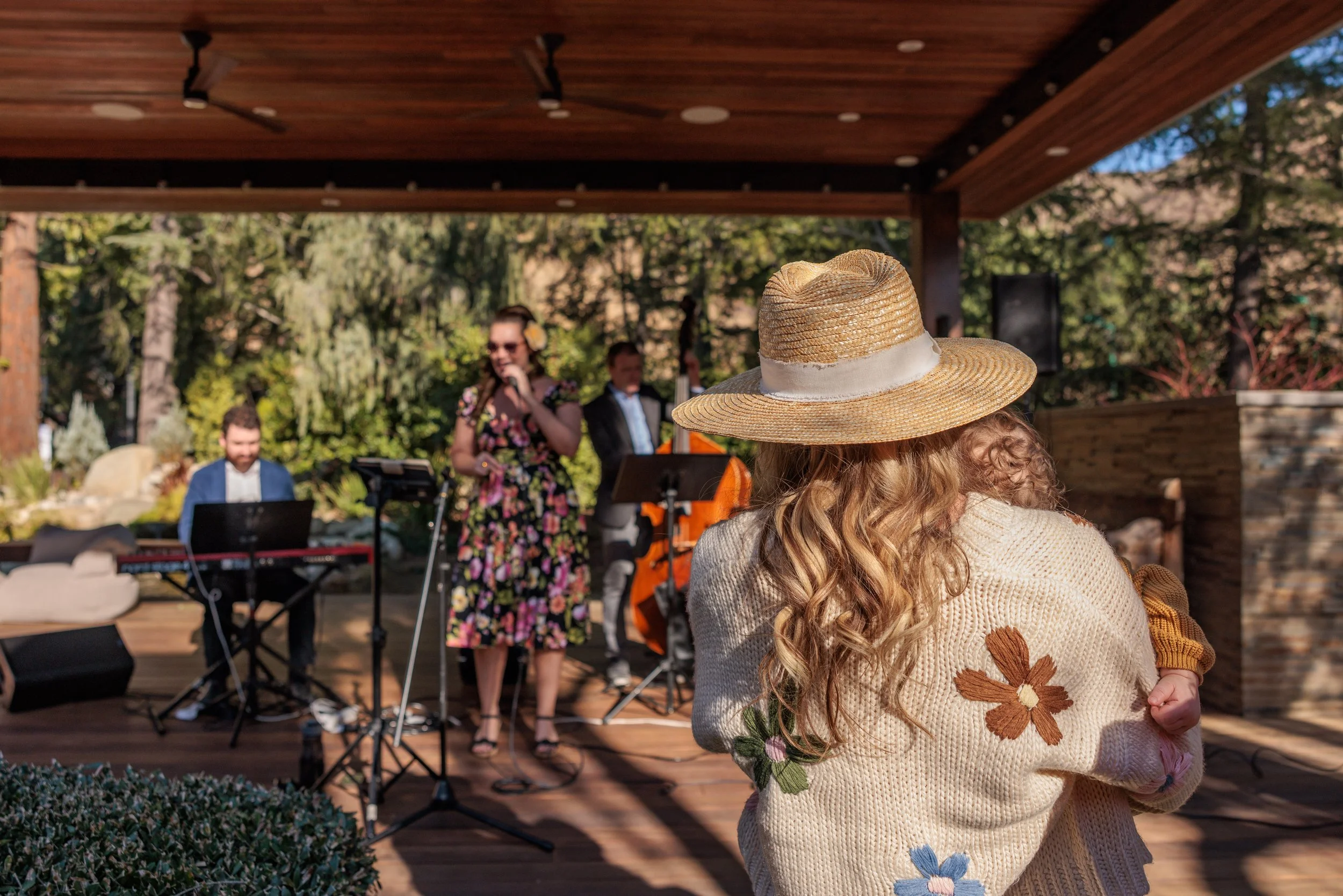 A woman with long curly hair wearing a sun hat and a sweater with embroidered flowers, holding a small child, watching a live outdoor music performance with a singer, keyboardist, and another musician in the background.