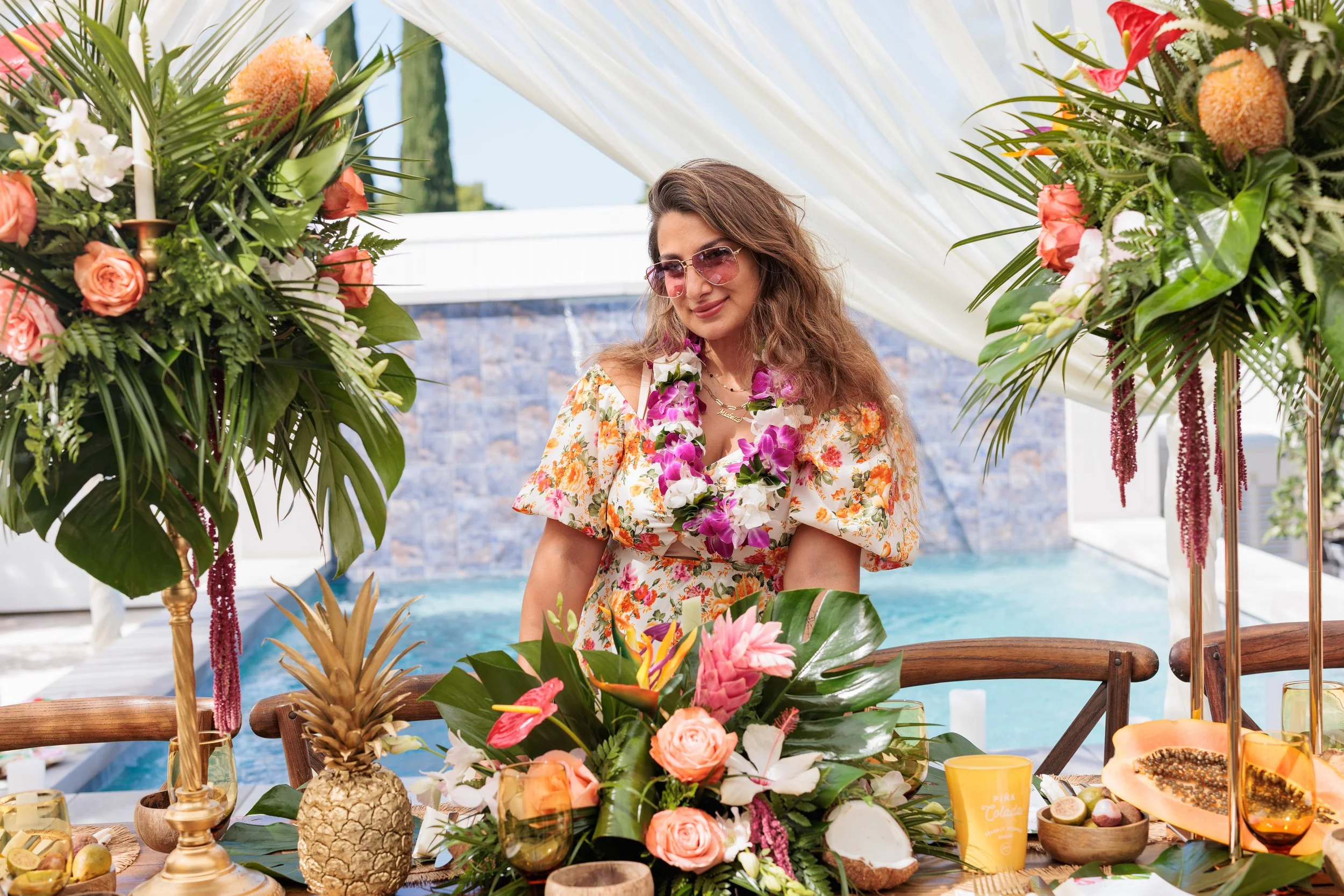 Woman in floral dress and sunglasses at a tropical-themed outdoor celebration with floral decorations, tropical fruits, and a pool in the background.