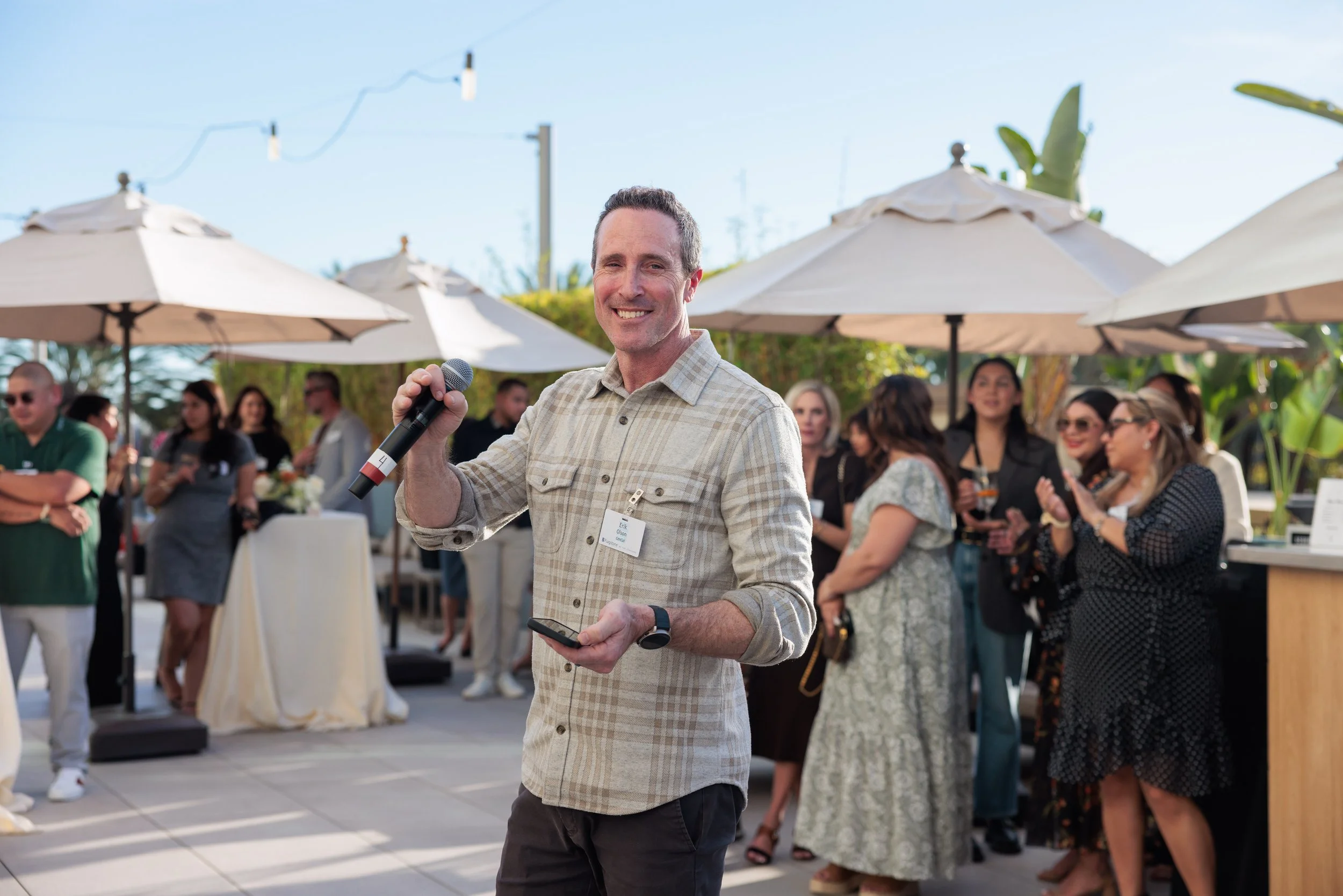 A man holding a microphone and a phone, smiling at a social outdoor gathering with food, drinks, and people under umbrellas.