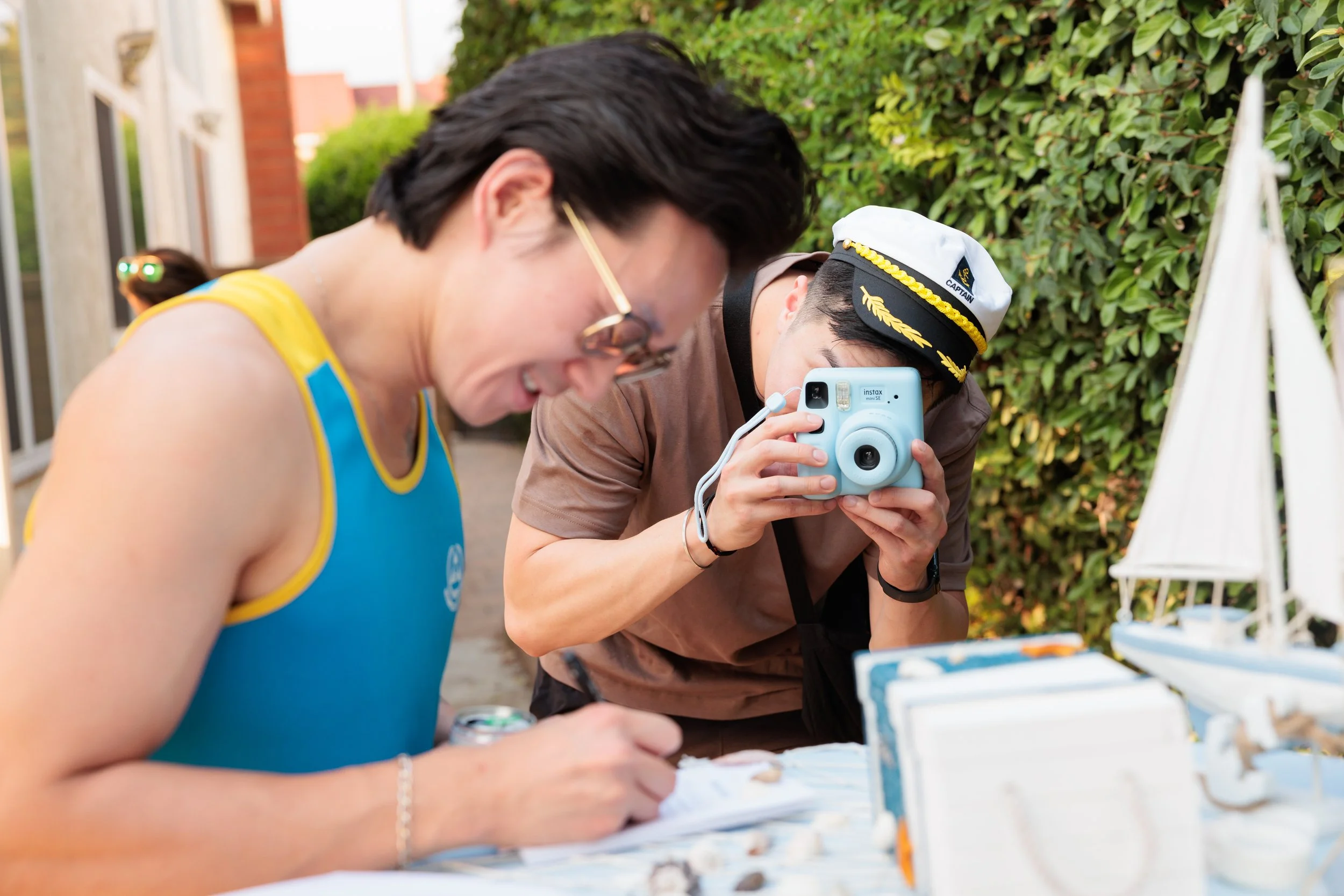 A person in a blue tank top with yellow trim is writing or drawing on paper at a table, while another person in a captain's hat and brown shirt is taking a photo with a light blue instant camera.