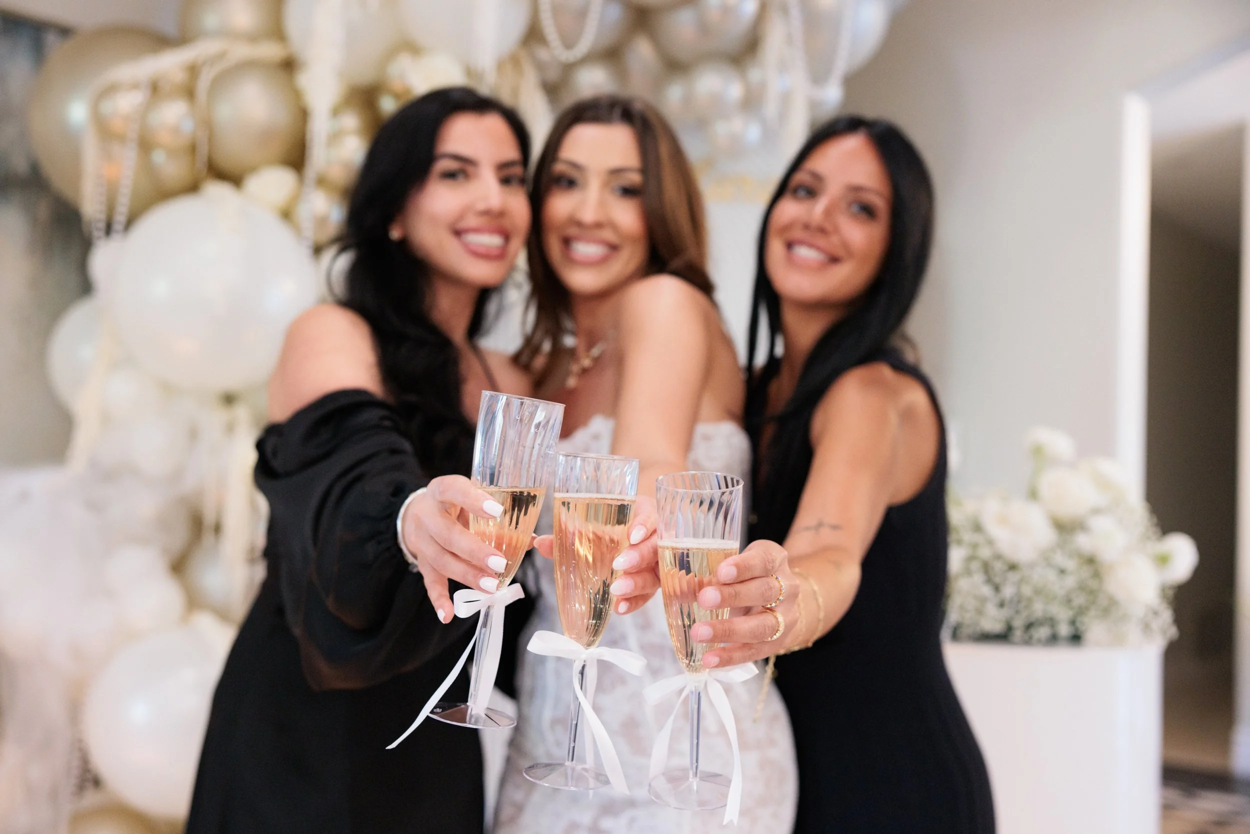 Three women celebrating with champagne glasses at a party or wedding reception with white balloon decorations in the background.