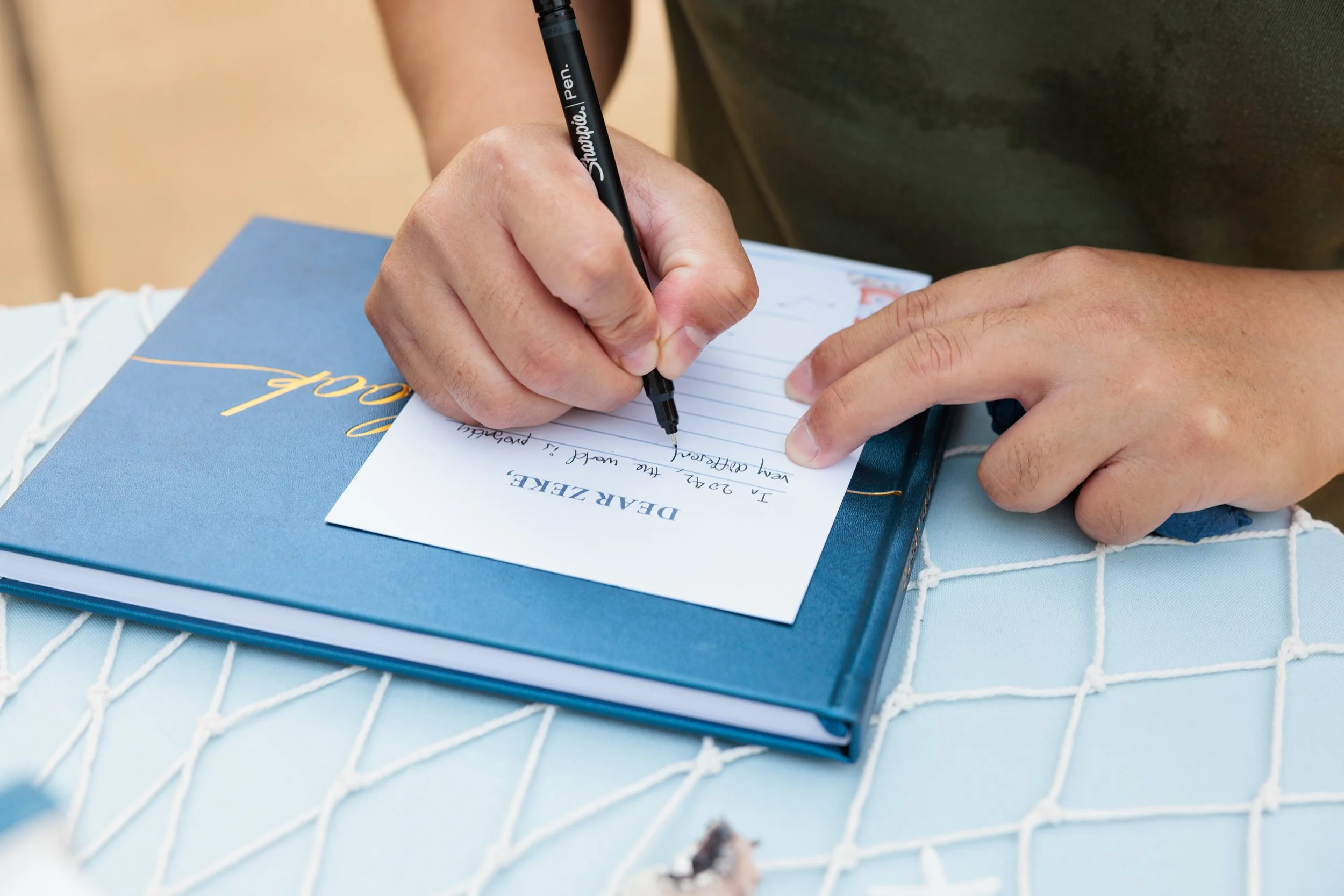 Person writing a farewell message on a card with a black Sharpie marker, with a blue folder or notebook underneath, sitting at a table covered with a white textured tablecloth.